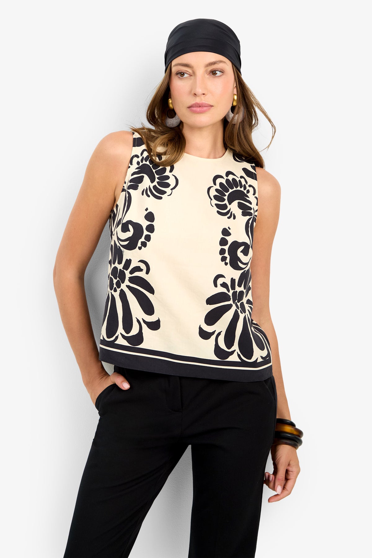 A woman models the MALEMA TOP—a sleeveless cream blouse with bold black floral patterns—paired with black pants, a black headscarf, gold hoop earrings, and black bangles, posing with one hand in her pocket against a light background.