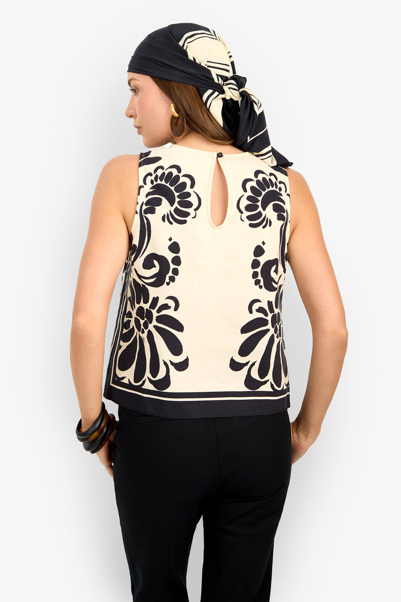 A woman, seen from behind, wears the MALEMA TOP—a sleeveless black and beige piece with bold floral back patterns—paired with black pants and a coordinating headscarf.