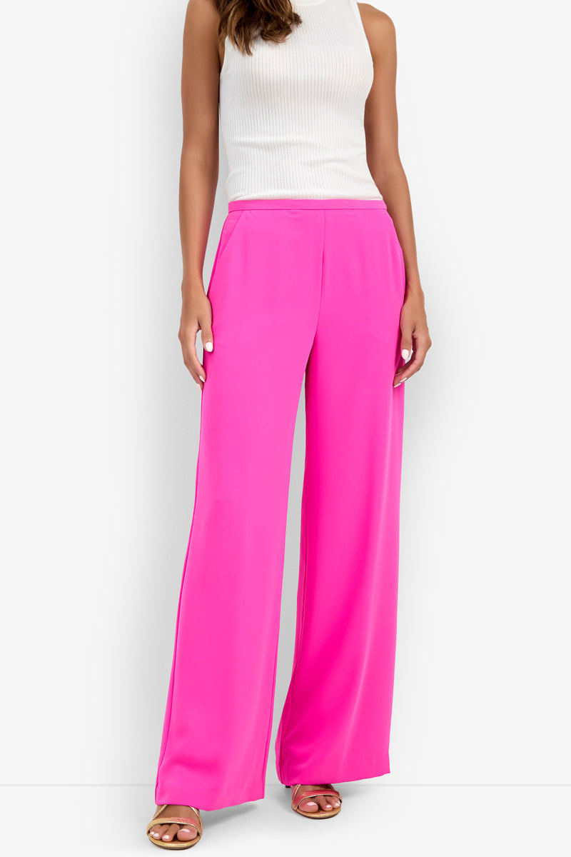 A woman is standing and facing forward against a plain white background, wearing the Lyndi Top, a white sleeveless ribbed top, paired with bright pink wide-leg pants and brown sandals.