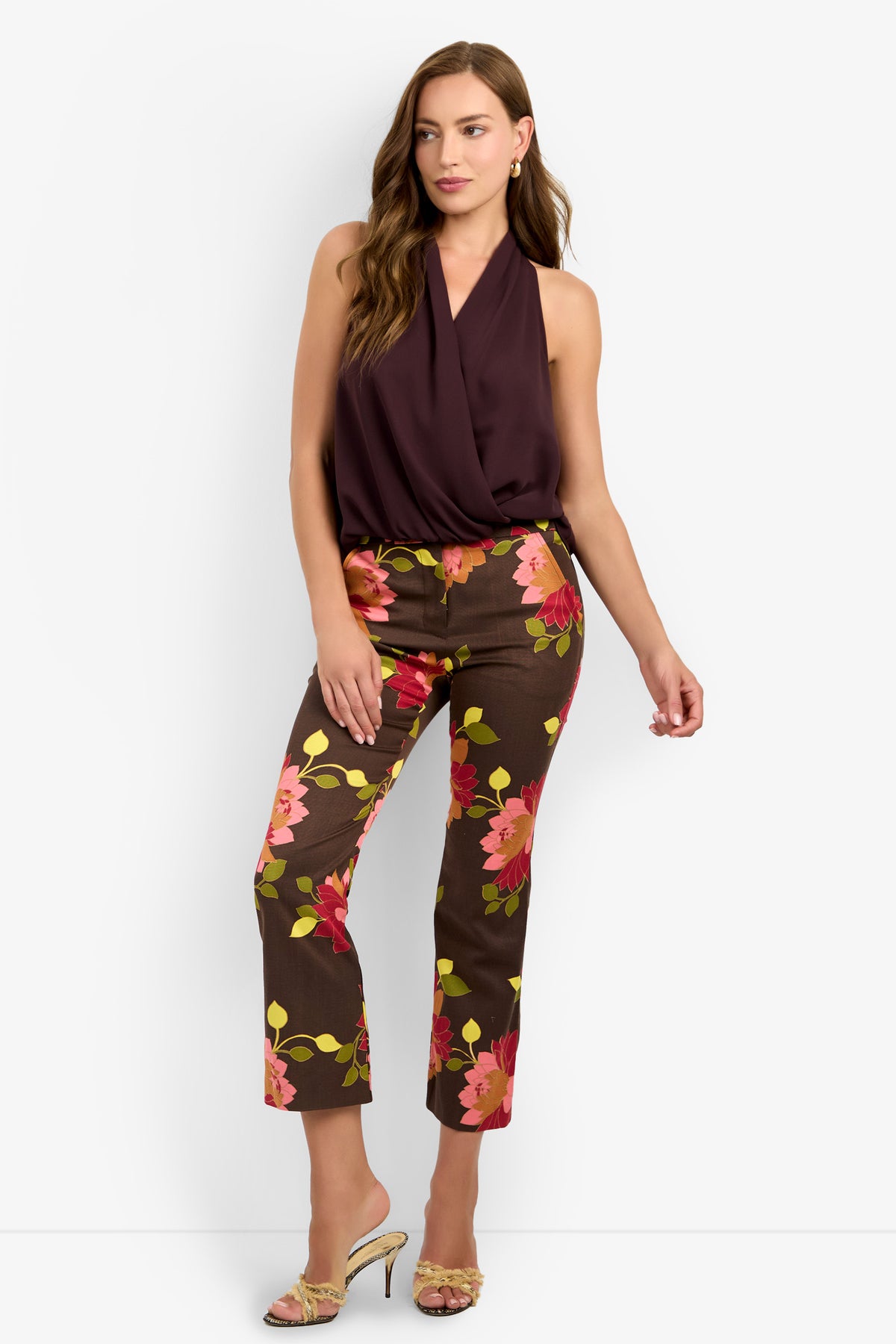 A woman stands against a white background wearing the LUCCA TOP, a sleeveless deep burgundy blouse, paired with brown pants featuring bold pink, orange, and yellow floral prints and open-toe heels.