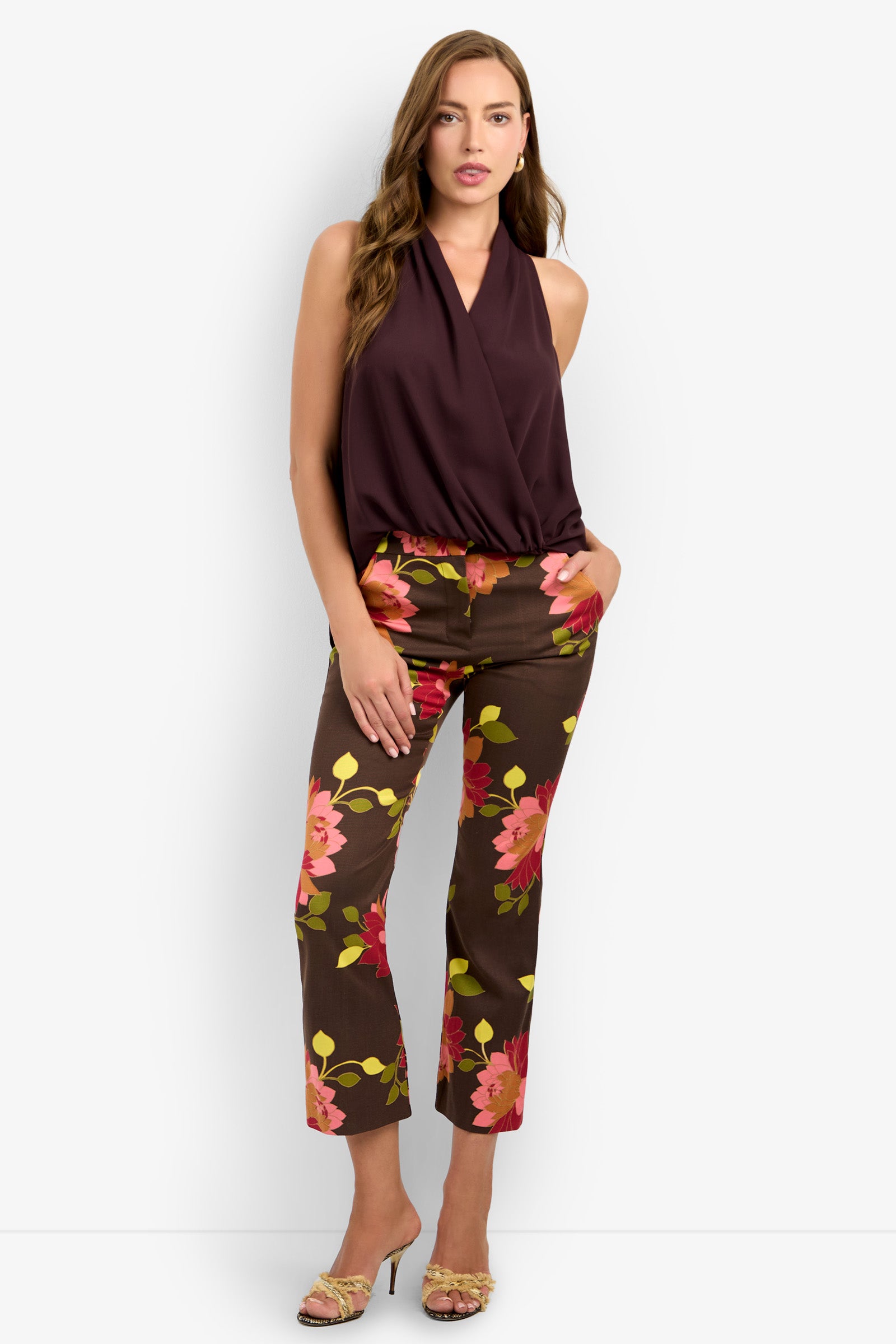 A woman stands against a white background wearing the LUCCA TOP, a sleeveless dark purple wrap top, paired with brown floral pants featuring pink and yellow flowers, and open-toe heels. She has long hair and a neutral expression.