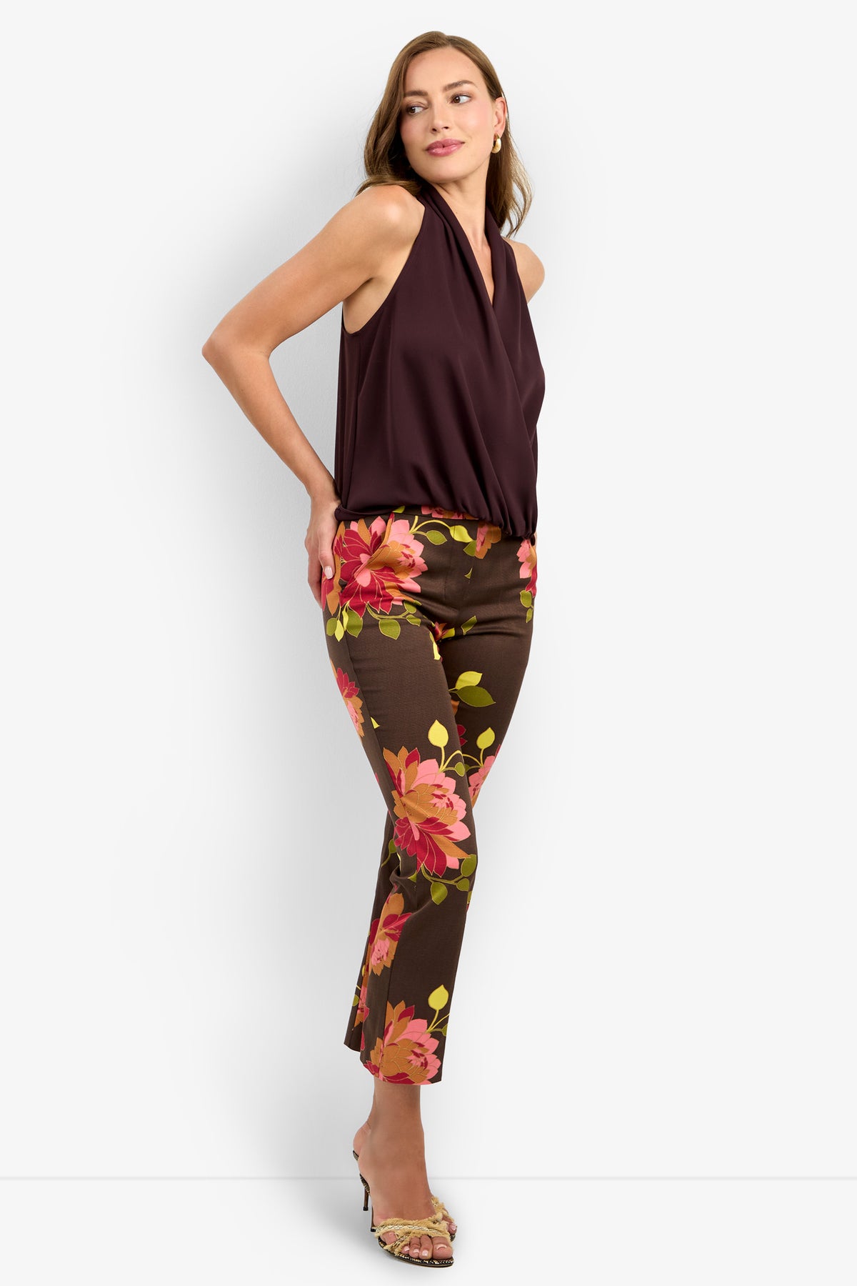 A woman poses in the sleeveless, deep burgundy LUCCA TOP styled with brown pants featuring bold pink and orange floral prints. She wears open-toe heels and her loose light brown hair completes the look.