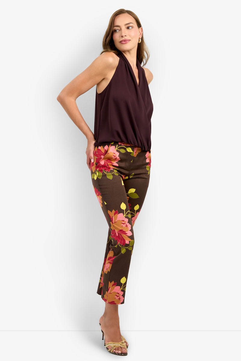 A woman poses in the sleeveless, deep burgundy LUCCA TOP styled with brown pants featuring bold pink and orange floral prints. She wears open-toe heels and her loose light brown hair completes the look.