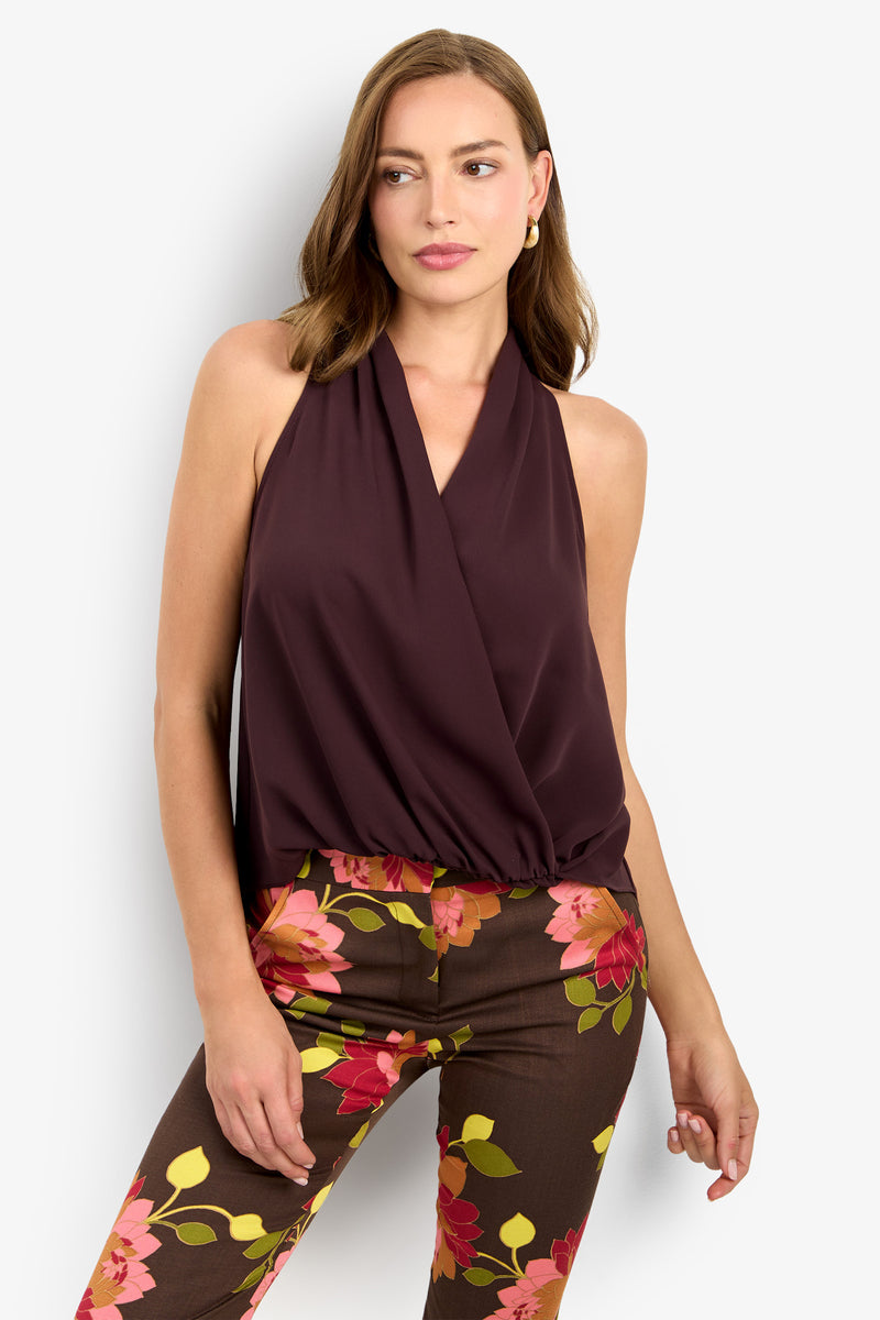A woman wears the LUCCA TOP, a sleeveless dark burgundy halter, paired with brown pants featuring a bold pink and orange floral pattern, standing against a plain white background.