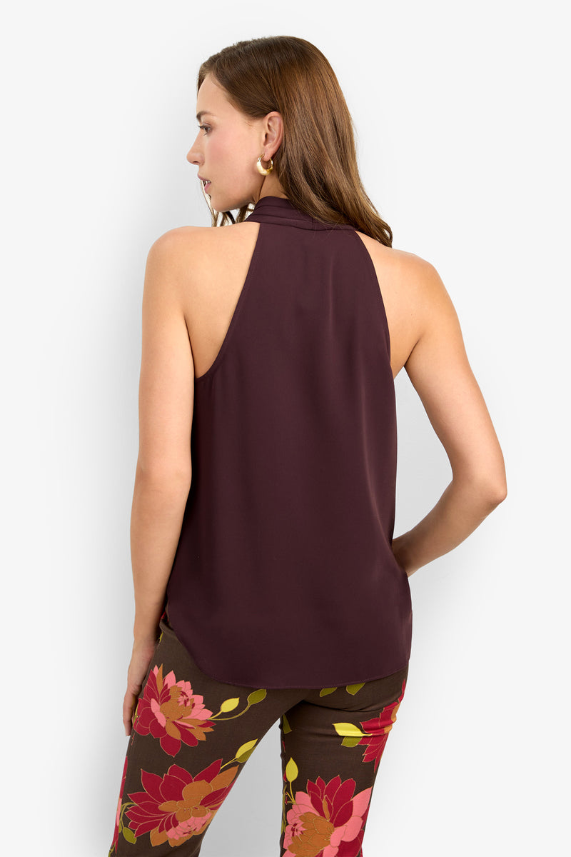 A woman with light brown hair wears the LUCCA TOP, a sleeveless halter-neck maroon top, paired with brown pants featuring bold red and yellow floral prints, standing with her back and head turned slightly to the side.