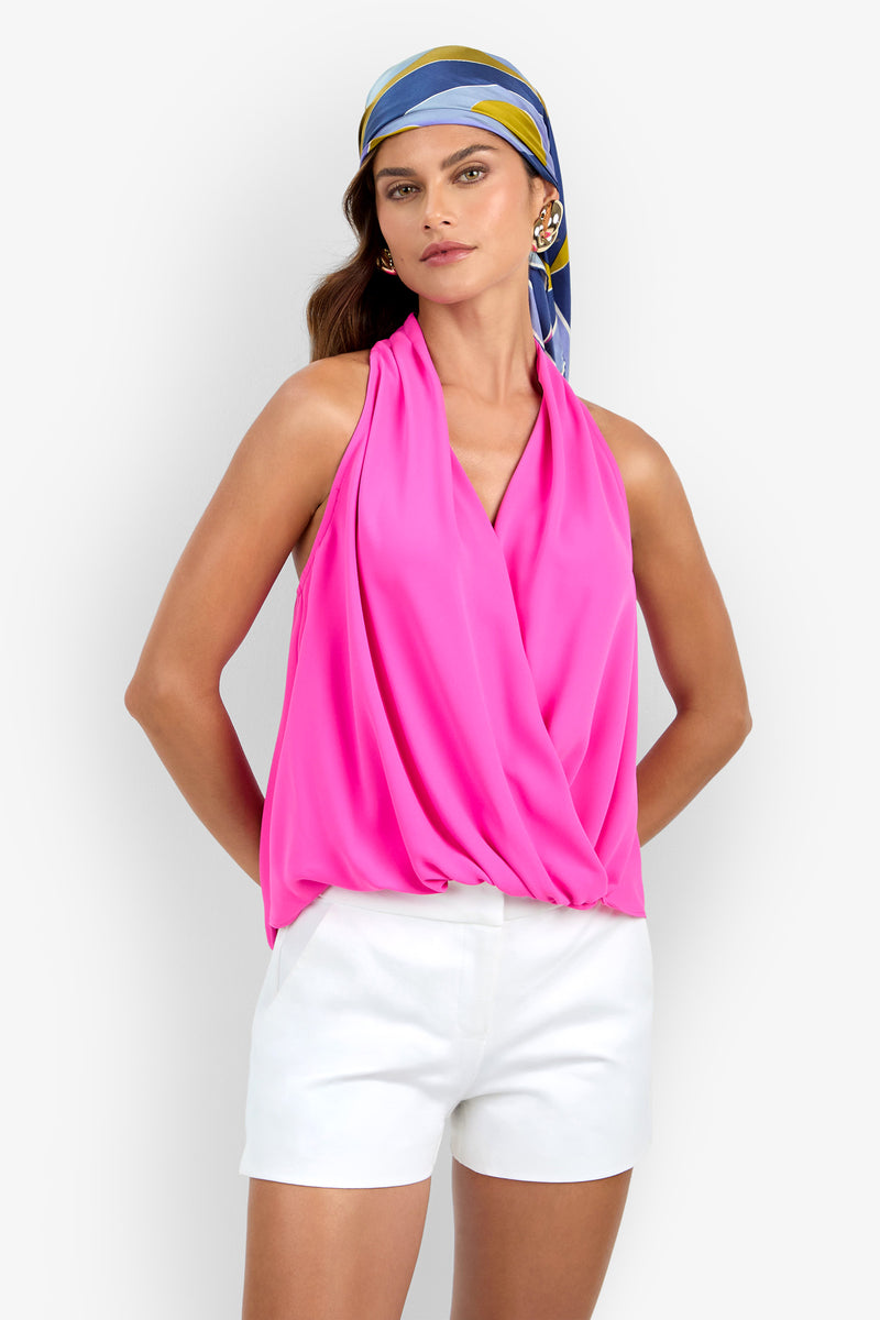 A woman is wearing the Lucca Top in bright pink, styled with white shorts, gold hoop earrings, and a blue, yellow, and white headscarf. She stands with her hands in her pockets against a plain background.