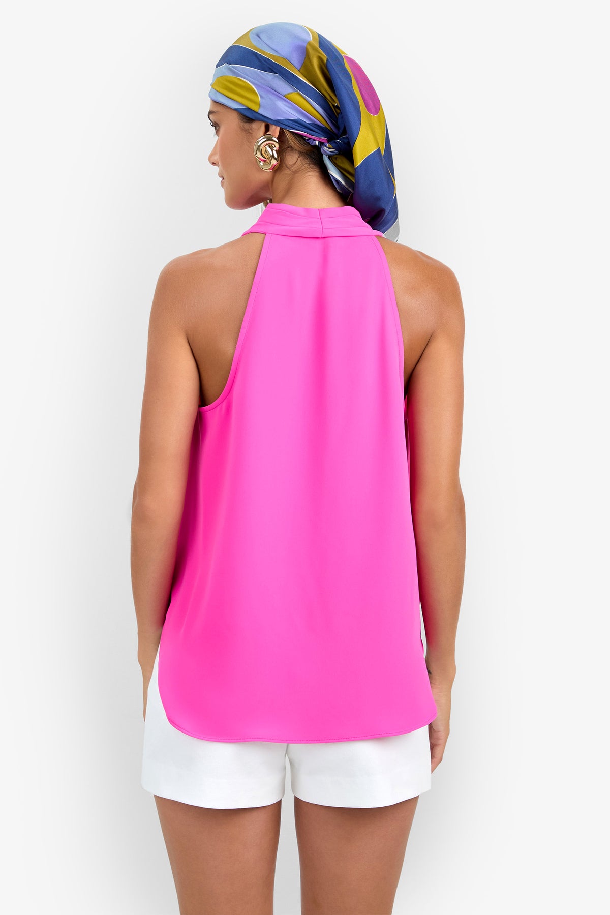 A woman facing away, wearing the bright pink Lucca Top, white shorts, and a colorful headscarf in shades of blue, yellow, and purple, stands against a plain light background.