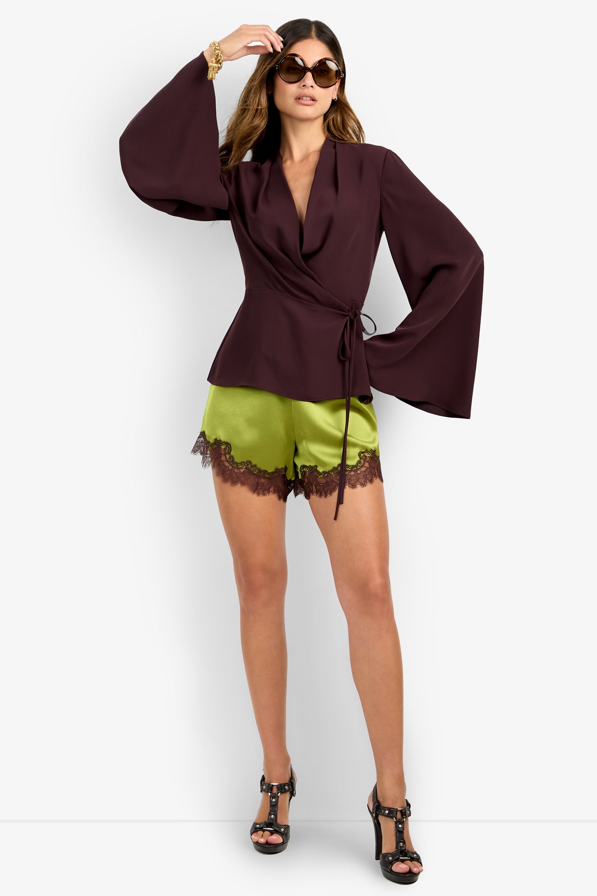 A woman poses confidently in the DEE DEE TOP, a dark purple wrap blouse with wide sleeves, styled with lime green satin lace-trim shorts, black high-heeled sandals, oversized sunglasses, and a gold bracelet.