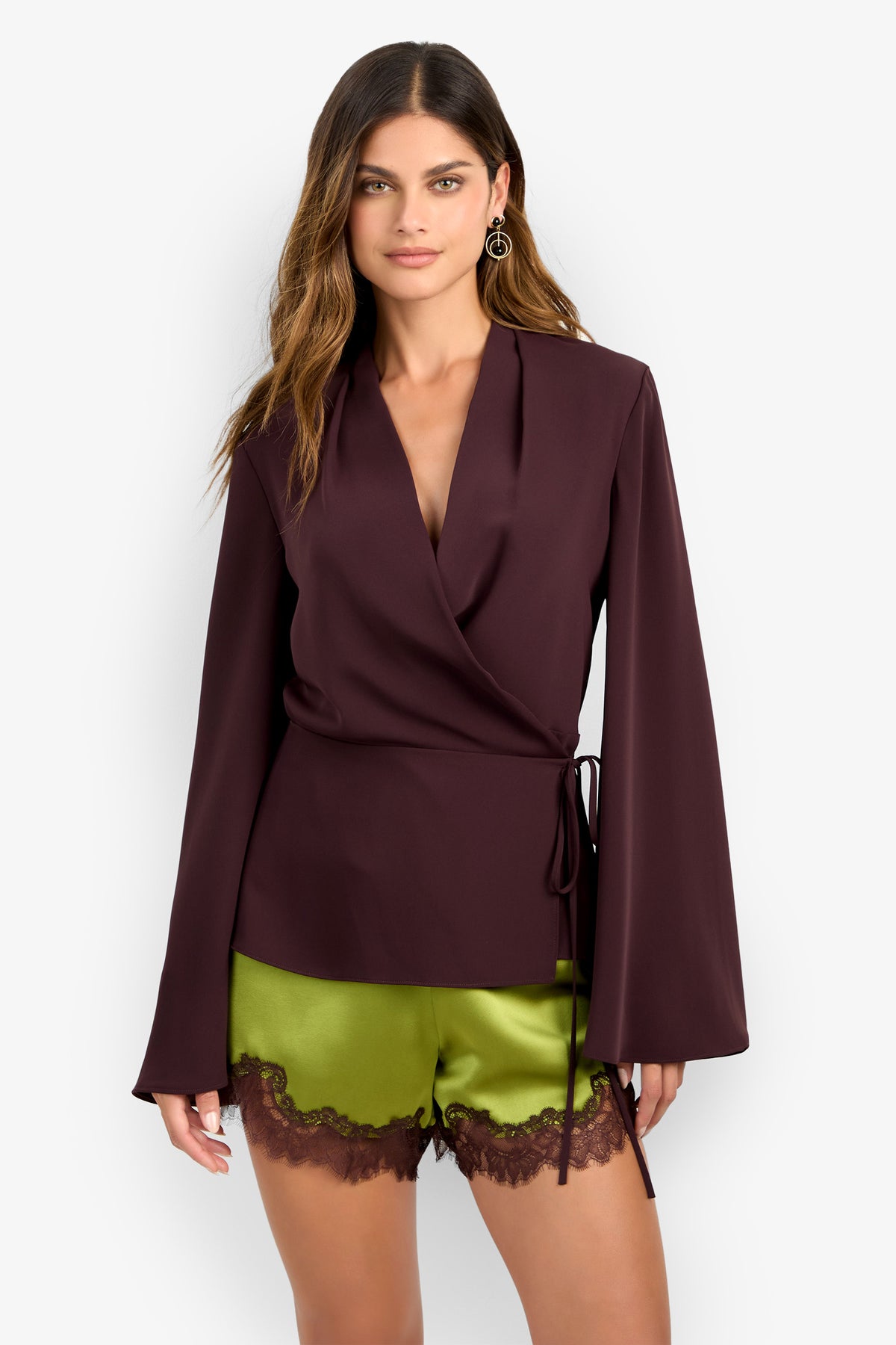 A woman wearing the DEE DEE TOP, a deep burgundy wrap blouse with long sleeves, and olive green satin shorts with brown lace trim, stands against a plain light background.