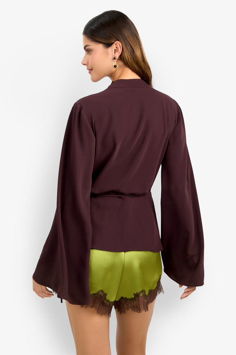A woman with long, wavy brown hair wears the DEE DEE TOP in burgundy with loose long sleeves and pairs it with lime green shorts trimmed in brown lace. She faces away and accessorizes with earrings.
