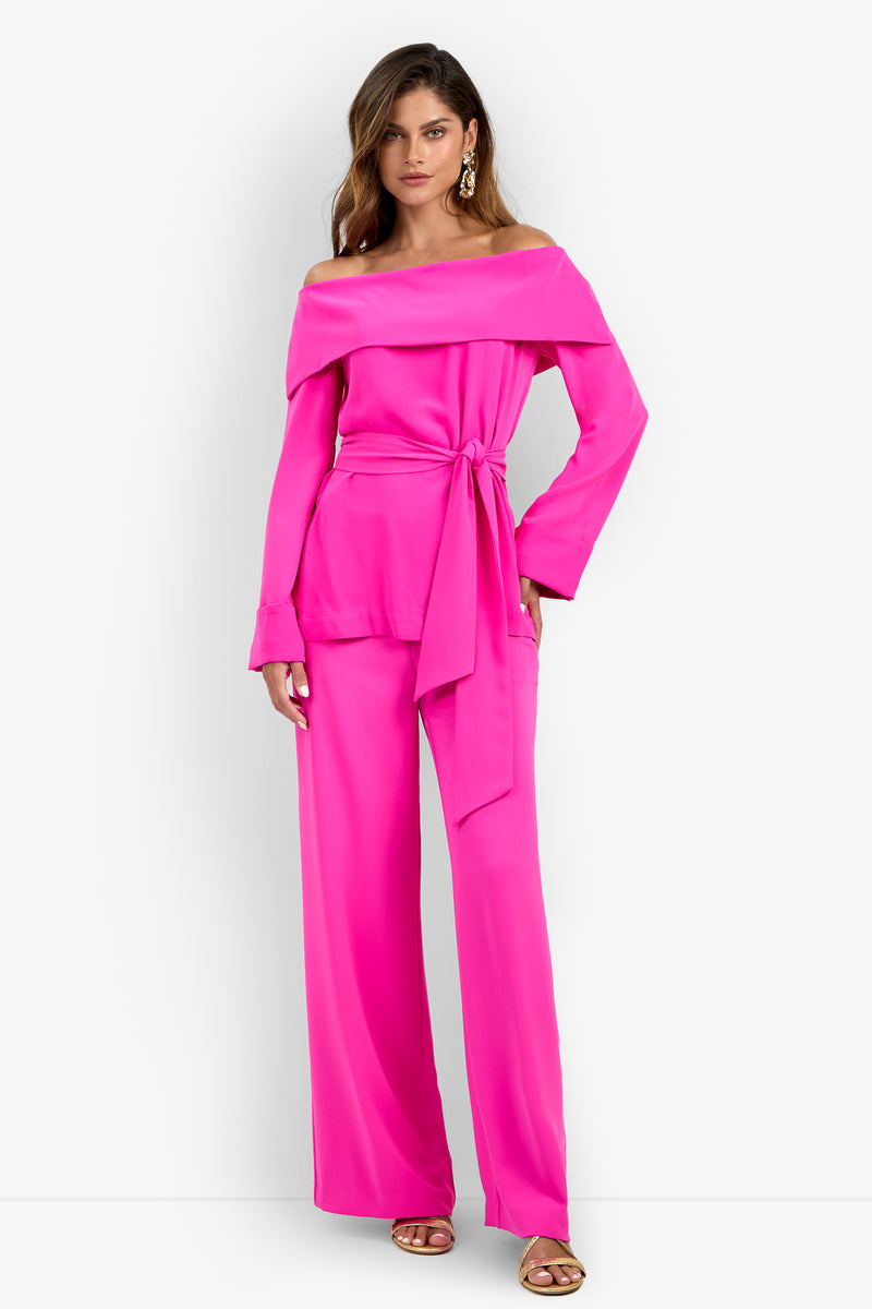 A woman is wearing the Lyndi Top in bright pink, styled off-the-shoulder with a matching sash belt and wide-leg pants. She stands against a white background, accessorized with gold earrings and sandals.