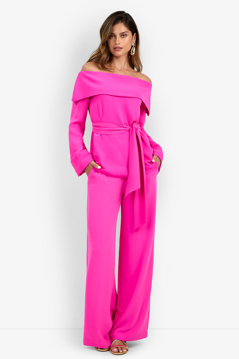 Woman wearing the Lyndi Top in a bright pink shade, styled off-the-shoulder with a belted waist and paired with wide-leg pants, standing against a plain white background with her hands in her pockets.