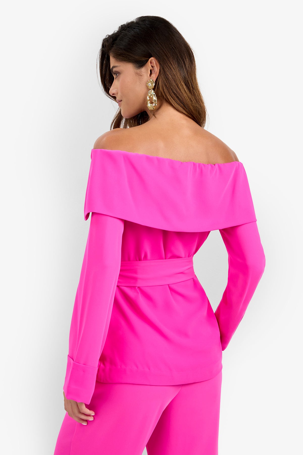 Woman facing away, wearing the bright pink Lyndi Top with a wide off-the-shoulder collar and matching pants. She has long brown hair, gold statement earrings, and her right hand on her hip against a white background.