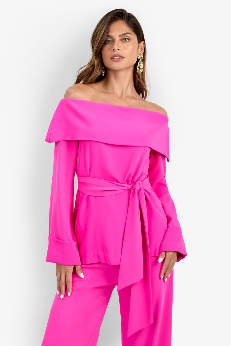 A woman models the Lyndi Top, a bright pink off-the-shoulder style with a wide collar, confidently posing with one hand on her hip against a plain white background.