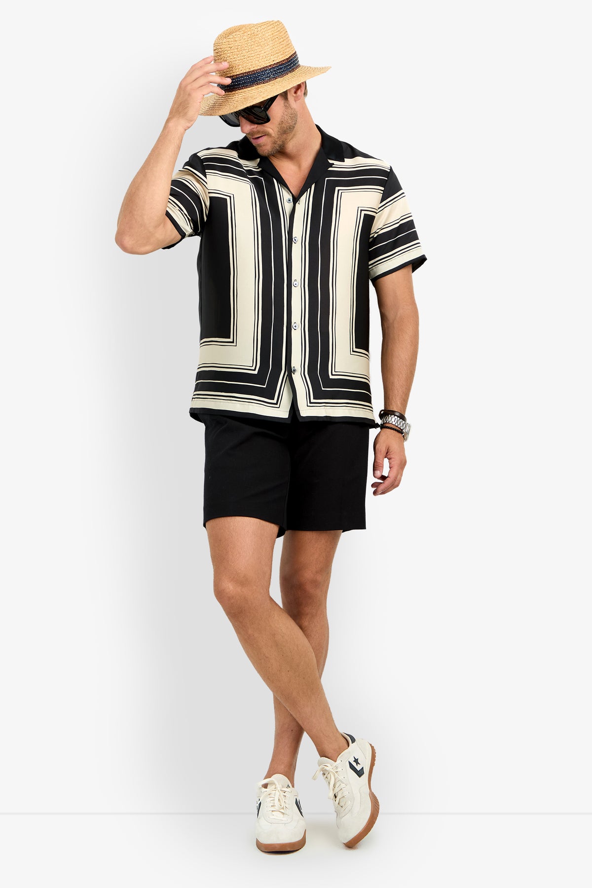 A man in a Felix Camp Shirt, black shorts, white sneakers, a straw hat, black sunglasses, and bracelets stands against a white background, touching his hat’s brim.
