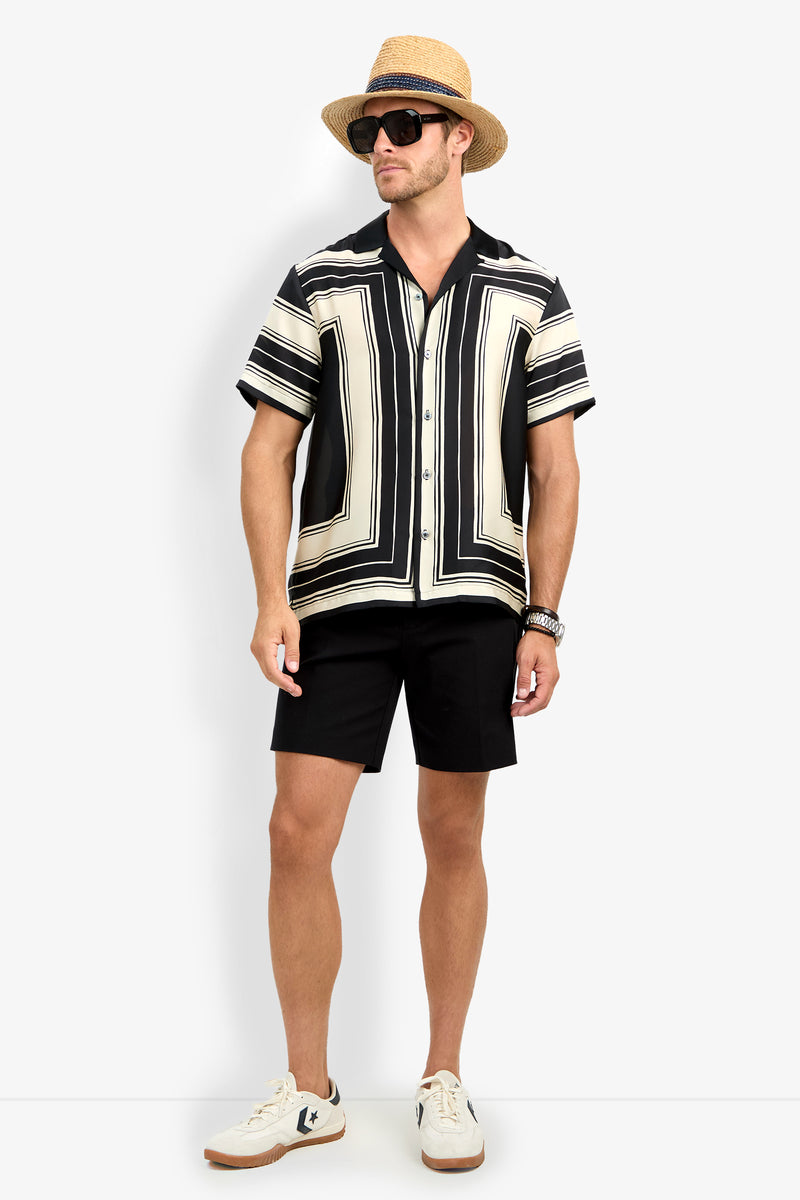 Man wearing a straw hat, black sunglasses, the Felix Camp Shirt (black and cream geometric pattern, short-sleeved), black shorts, white sneakers, and a watch, standing against a plain white background.