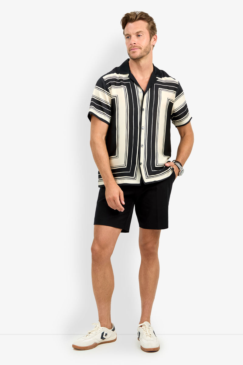 A man stands against a plain light gray background wearing the Felix Camp Shirt, which features black and cream geometric patterns, paired with black shorts, white sneakers with navy accents, and a watch.
