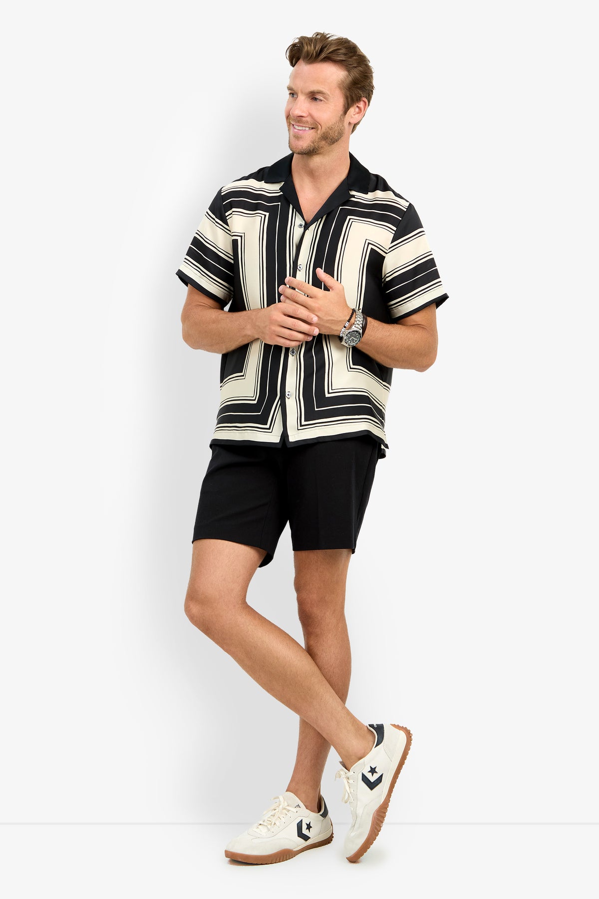 A man stands smiling, wearing the Felix Camp Shirt with a black and white geometric pattern, black shorts, white sneakers, and a watch; his arms are slightly raised and his legs crossed at the ankles.