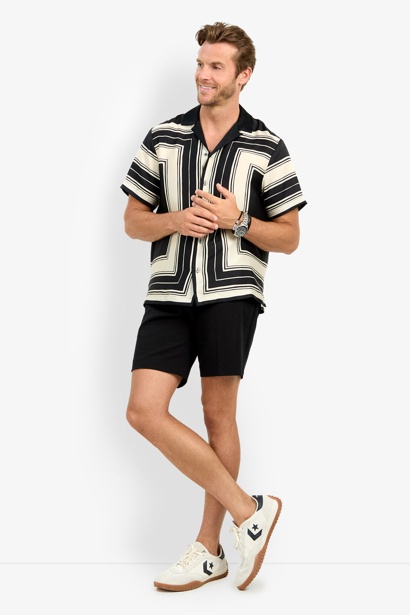 A man stands smiling, wearing the Felix Camp Shirt with a black and white geometric pattern, black shorts, white sneakers, and a watch; his arms are slightly raised and his legs crossed at the ankles.