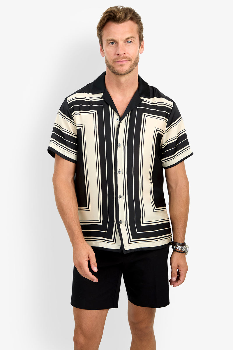 A man stands against a plain white background wearing the Felix Camp Shirt, a short-sleeve button-up with a bold black and cream geometric pattern, paired with black shorts and a watch on his left wrist.