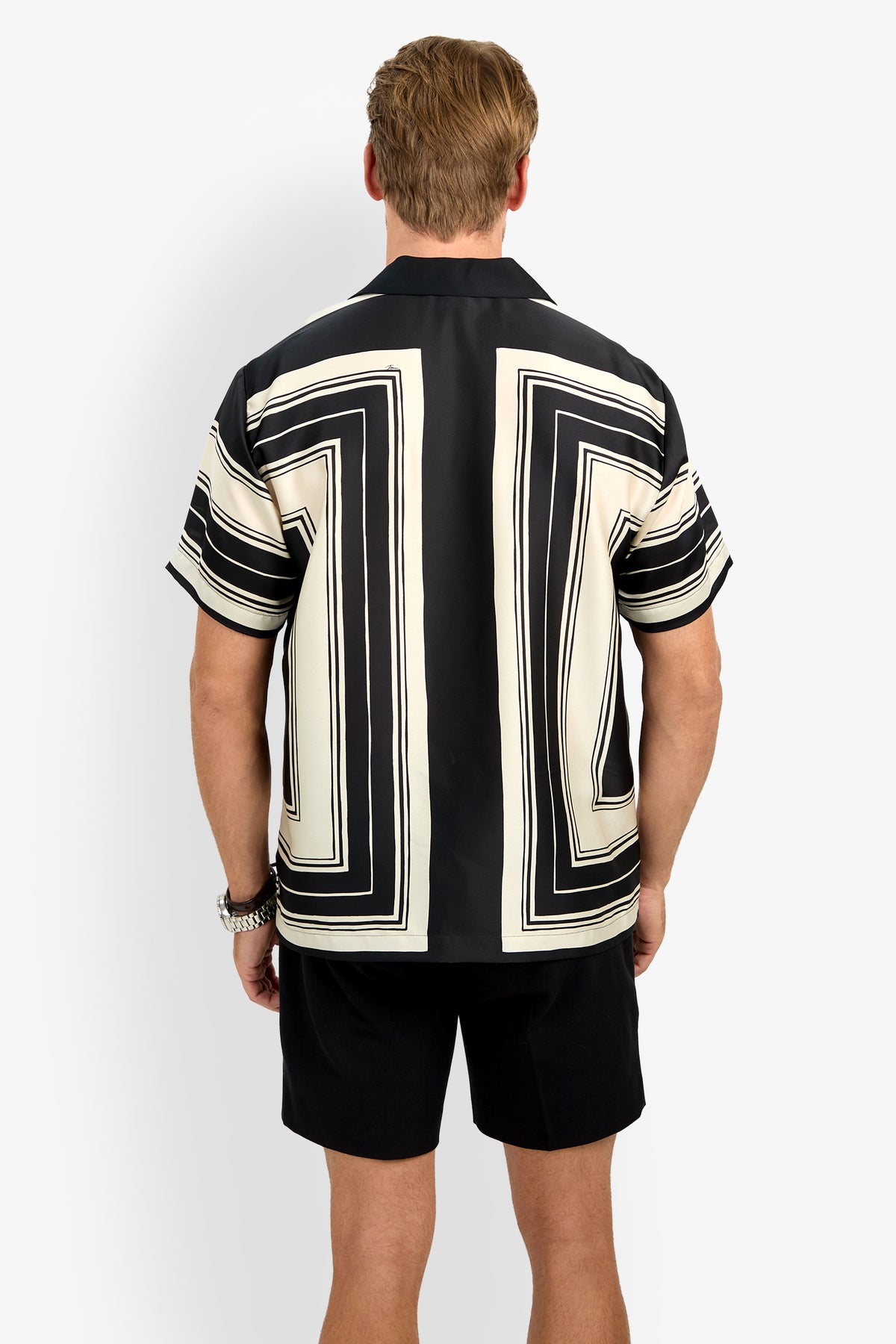A man stands facing away from the camera, wearing the Felix Camp Shirt, a short-sleeve top with a bold black and white geometric pattern featuring large rectangular designs on the back and sleeves, paired with black shorts.