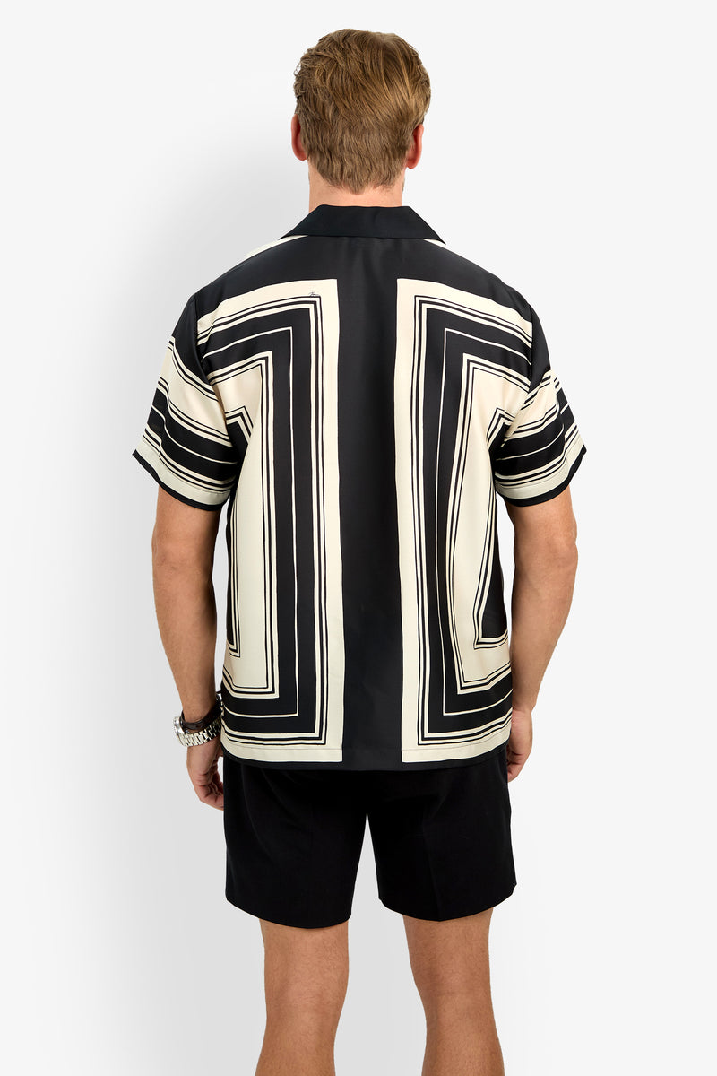 A man stands facing away from the camera, wearing the Felix Camp Shirt, a short-sleeve top with a bold black and white geometric pattern featuring large rectangular designs on the back and sleeves, paired with black shorts.