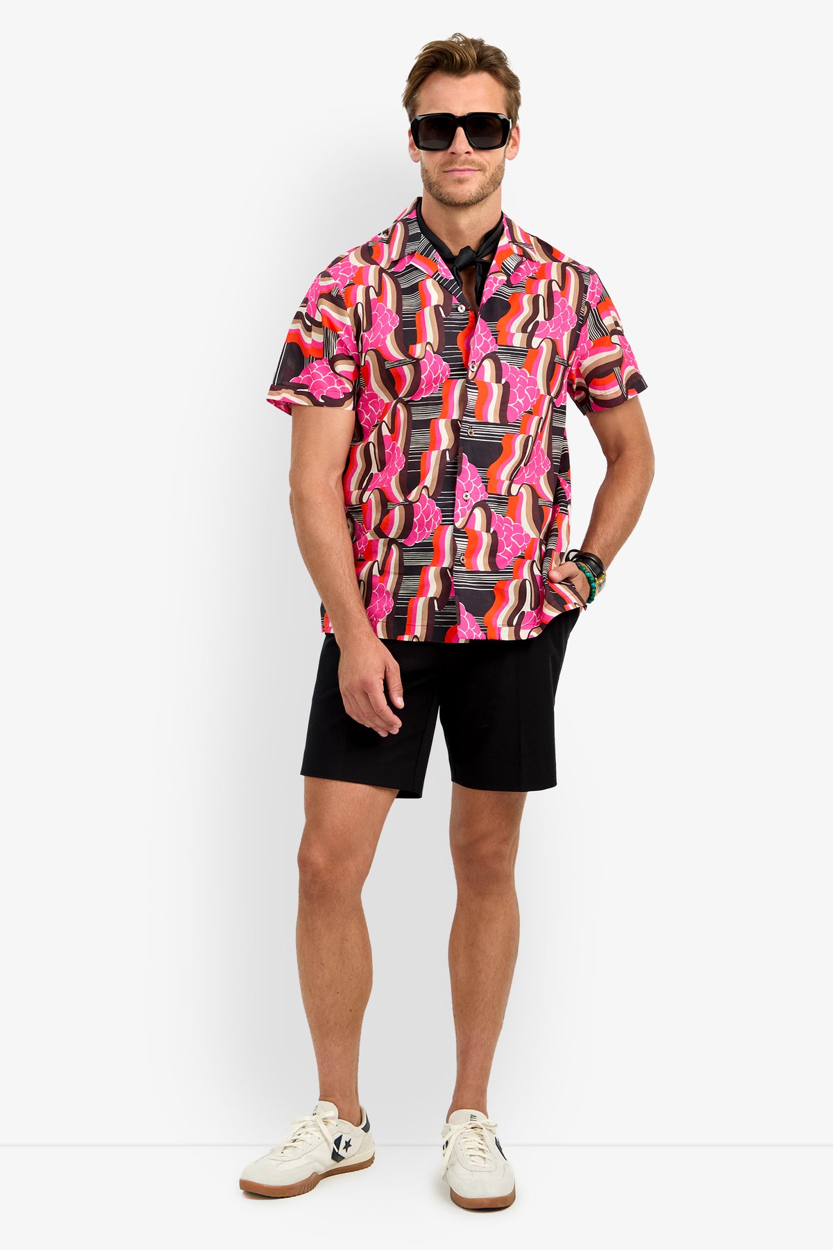 A man stands confidently against a plain white background wearing black sunglasses, the FELIX CAMP SHIRT in Giostra, black shorts, white sneakers, and bracelets.