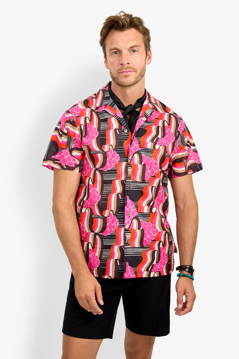 A man stands against a white background wearing the FELIX CAMP SHIRT, a colorful short-sleeve button-up with a bold pink, red, orange, and black abstract pattern, paired with black shorts and a black neck scarf.