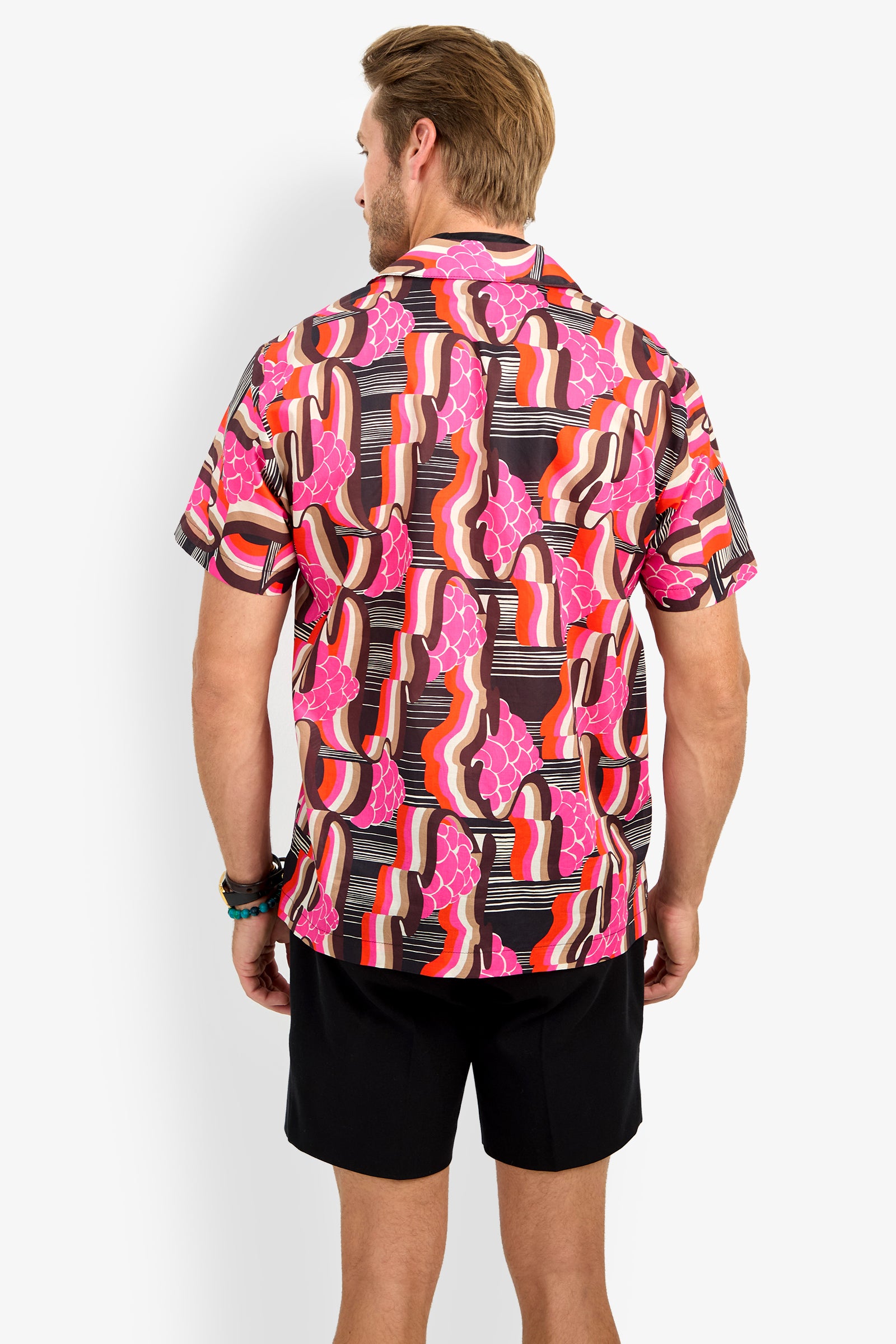 A man faces away, wearing the FELIX CAMP SHIRT—a colorful short-sleeve button-up with a bold abstract pattern in pink, orange, red, black, and white—paired with black shorts.