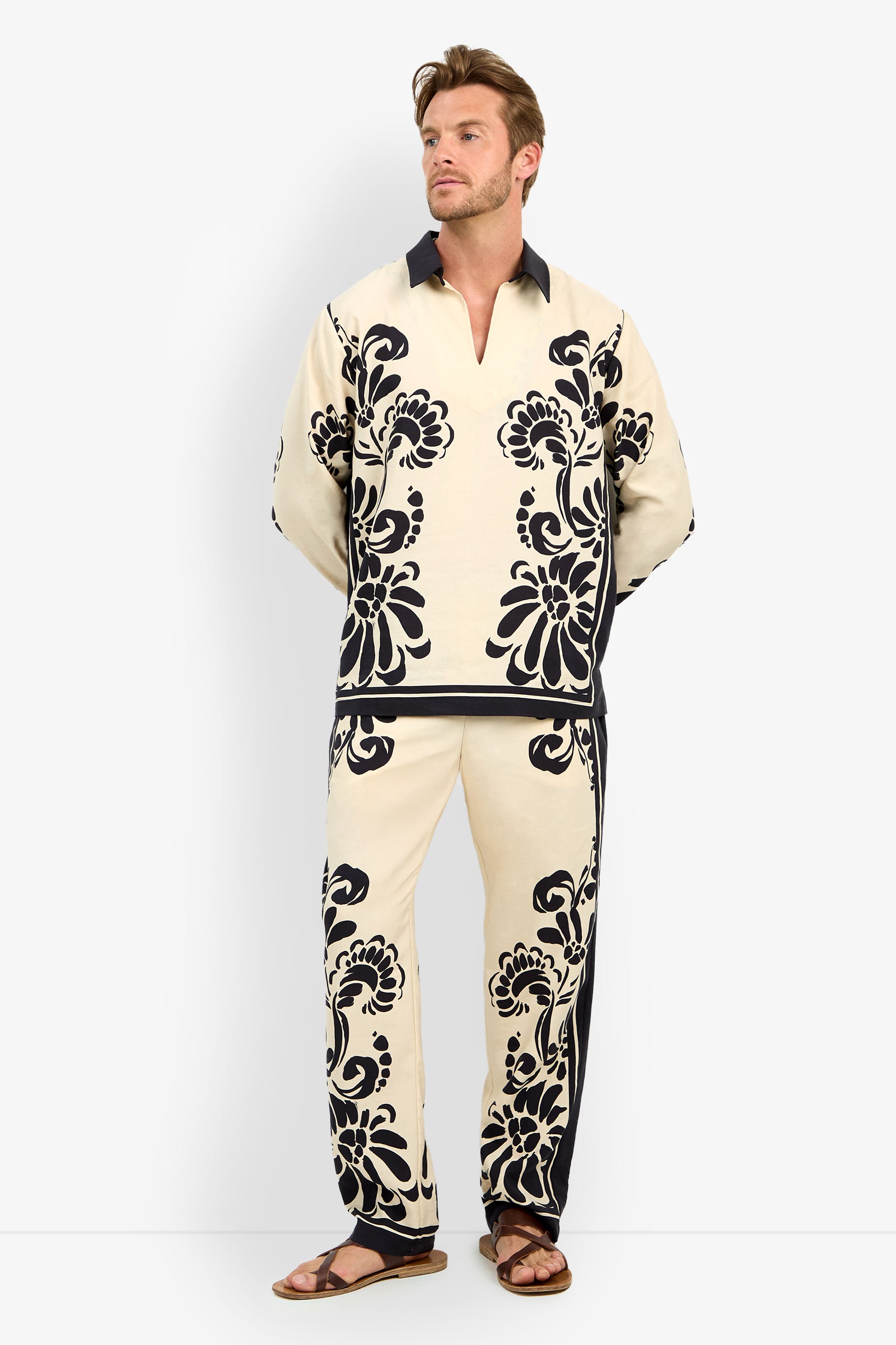 A man with light brown hair and a short beard wears the HOLMES PULLOVER, a cream-colored shirt featuring bold black floral patterns, paired with matching pants and brown sandals, standing against a plain white background.