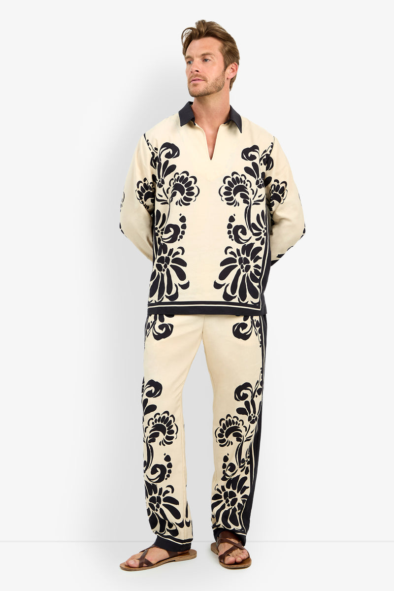 A man with light brown hair and a short beard wears the HOLMES PULLOVER, a cream-colored shirt featuring bold black floral patterns, paired with matching pants and brown sandals, standing against a plain white background.