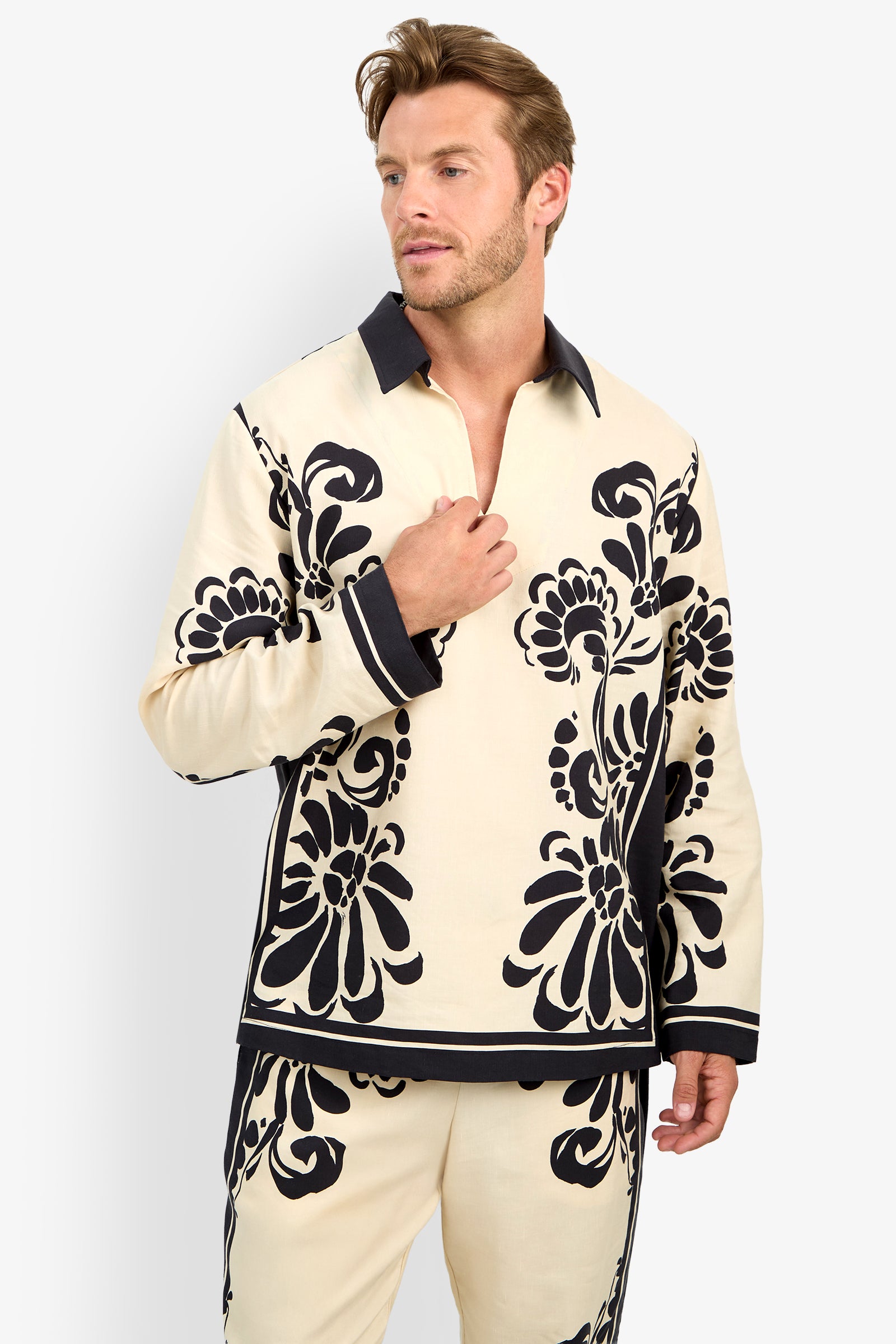 A man models the HOLMES PULLOVER, a cream-colored top with bold black floral patterns, dark collar and cuffs, paired with matching pants, standing against a plain white background.