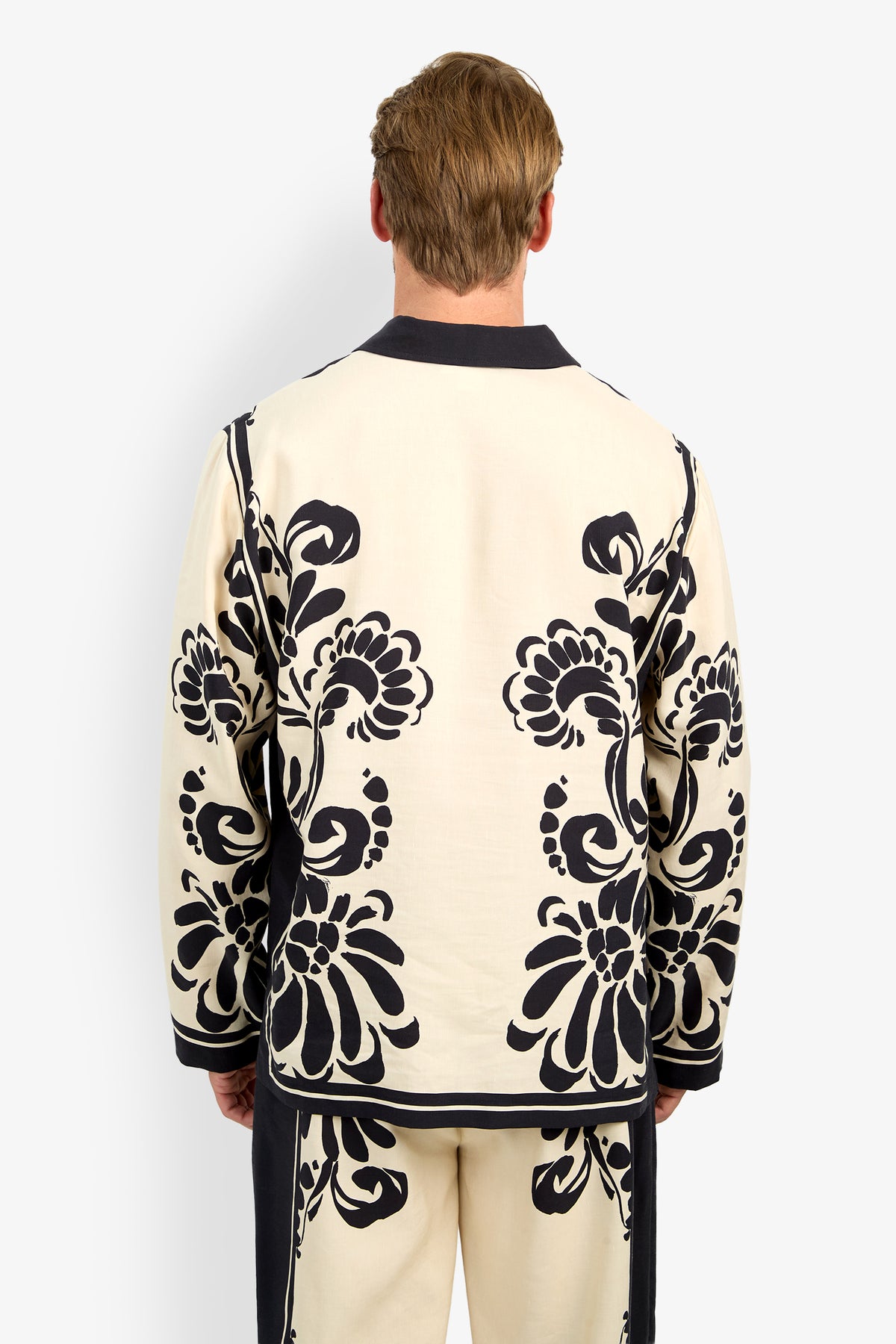 A person with light brown hair, seen from behind, is wearing the HOLMES PULLOVER—a cream piece featuring bold black floral patterns. The plain white background emphasizes the pullover’s striking design.
