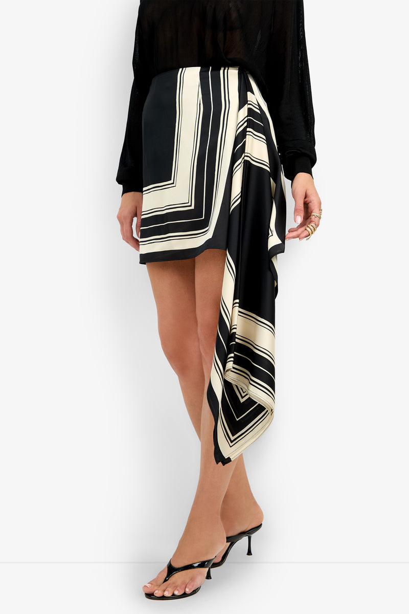 A woman wearing a black long-sleeve top, the FLORELLA SKIRT—a black and cream geometric asymmetrical mini skirt with draped detail—heeled sandals, and gold rings.