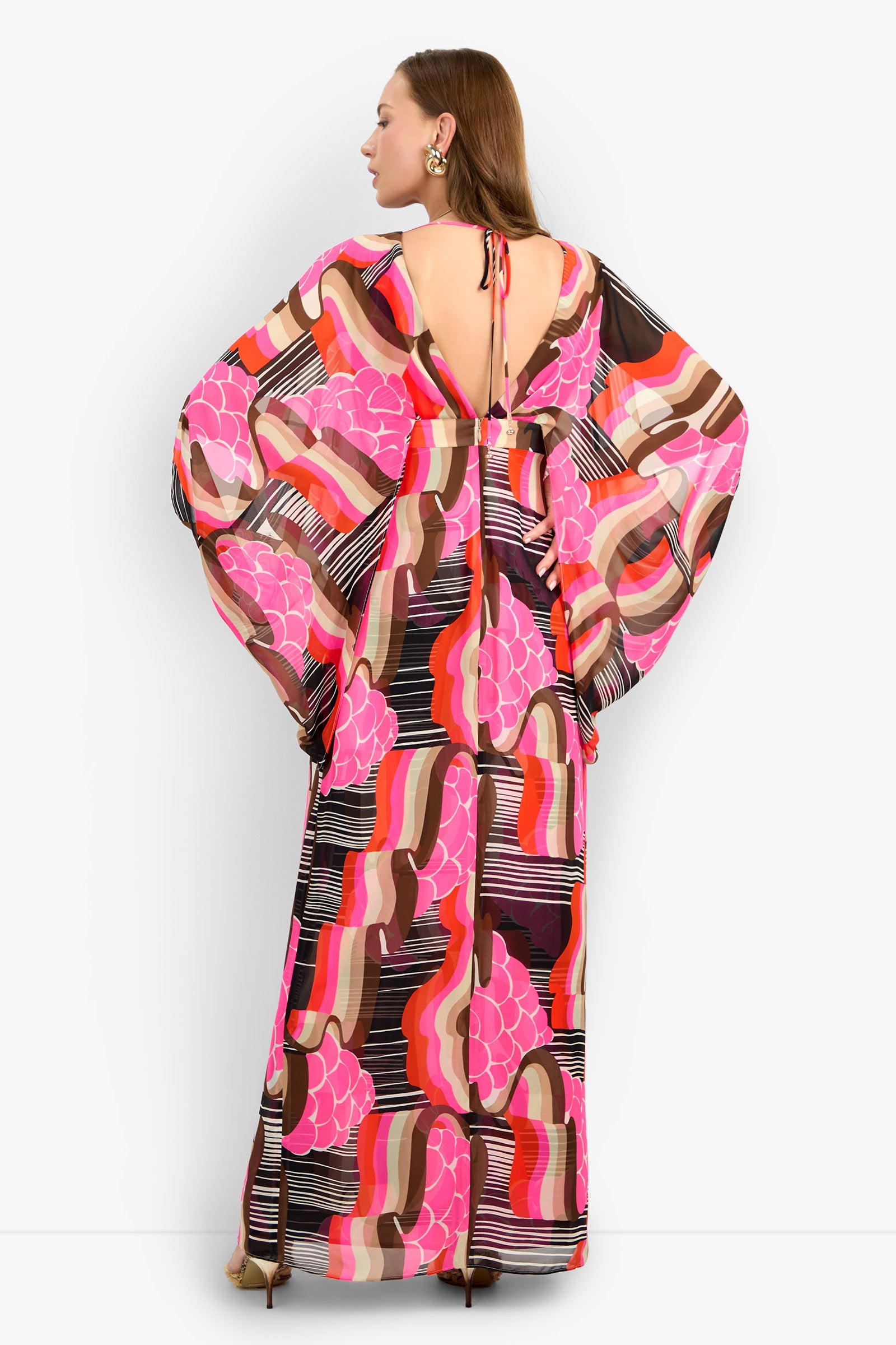 A woman is shown from the back wearing the BLOSSOM DRESS, a long, flowy dress with wide sleeves and a pink, red, brown, and white geometric pattern. It features a low back and a tie at the neck.