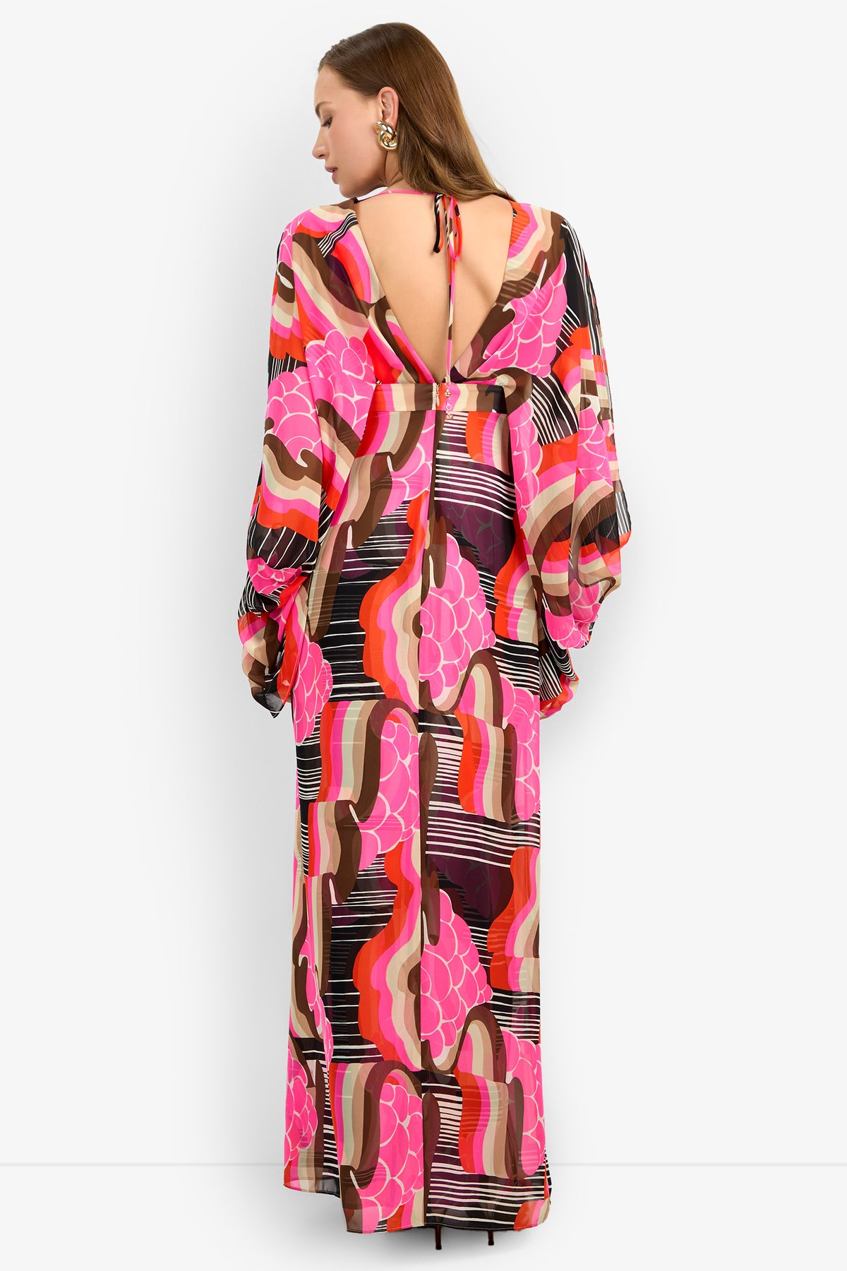 Woman facing away in the BLOSSOM DRESS, a long flowy dress with vibrant pink, brown, orange, and black geometric and floral patterns. Features an open back with neck tie and long voluminous sleeves.