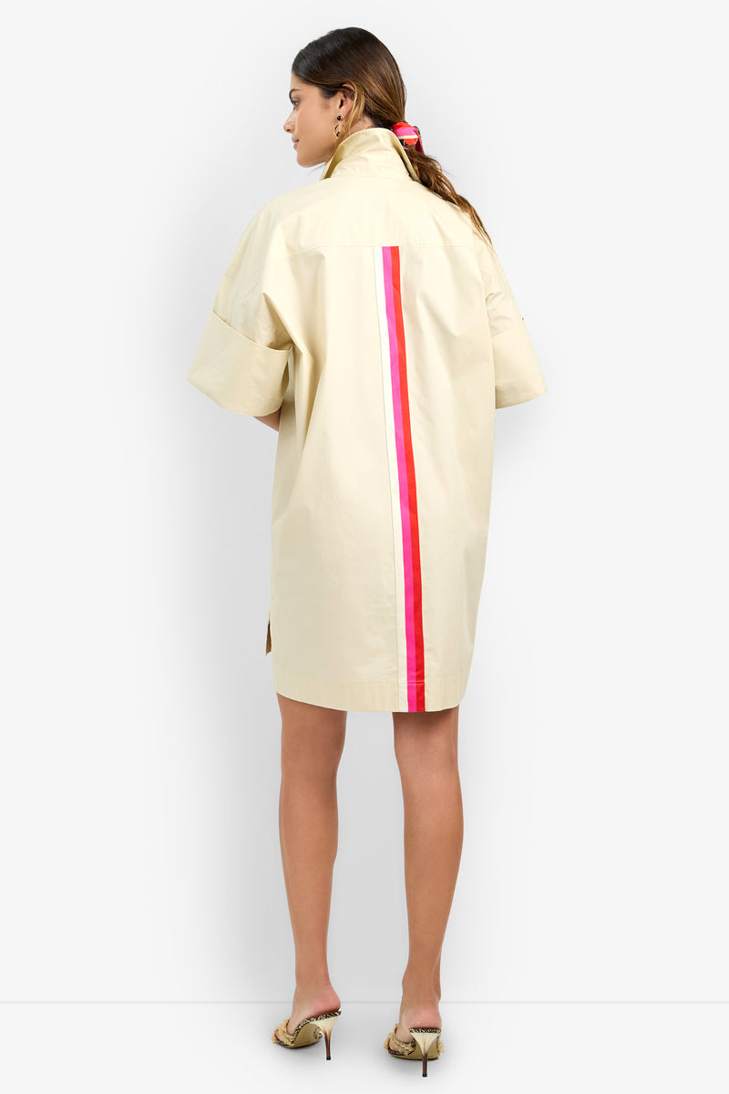 A woman with long hair faces away, wearing the OPPONENT DRESS—a loose, short-sleeve beige shirt dress featuring vertical red and pink stripes down the back. She pairs it with heeled sandals.