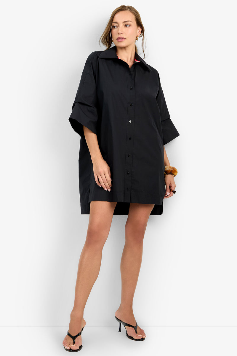 A woman stands against a white background wearing the loose-fitting black OPPONENT DRESS, black heeled sandals, and a brown hair scrunchie on her wrist.
