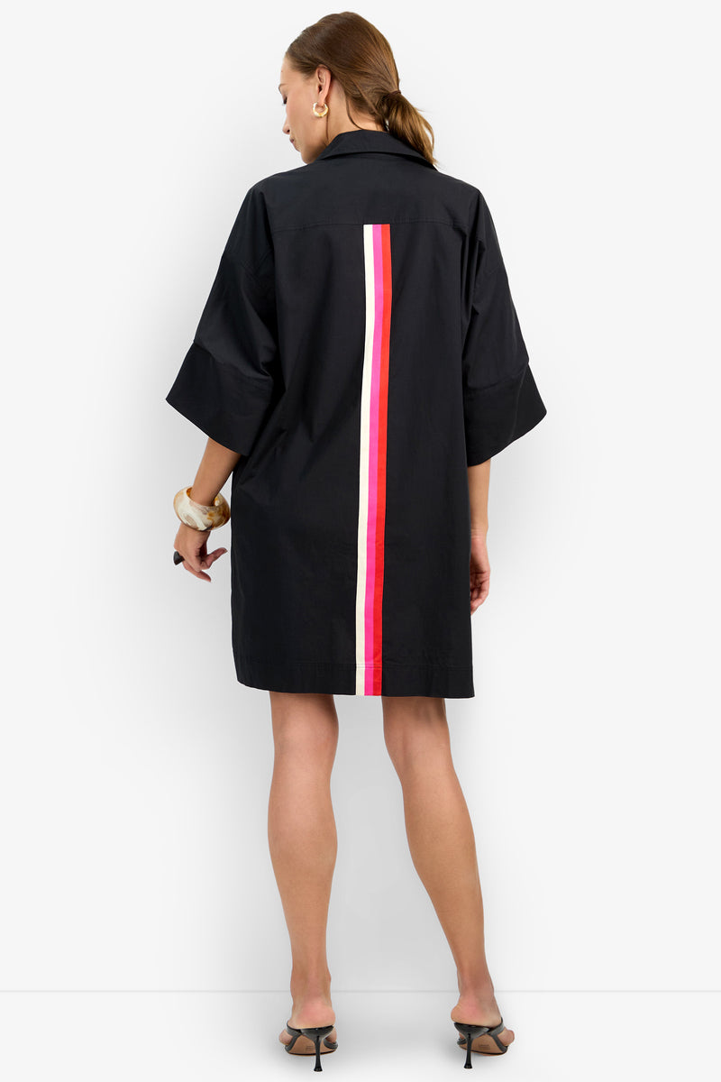 A woman with a ponytail faces away, wearing the OPPONENT DRESS—a loose black shirt dress with wide sleeves and a vertical red and white back stripe. She styles it with black heeled sandals and chunky bracelets.