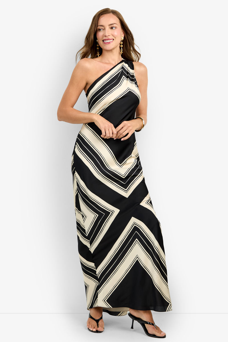 Woman wearing the CHIANTI DRESS, a black and cream one-shoulder maxi with bold geometric stripes, paired with black heeled sandals, large gold earrings, and a bracelet, smiles against a plain white background.