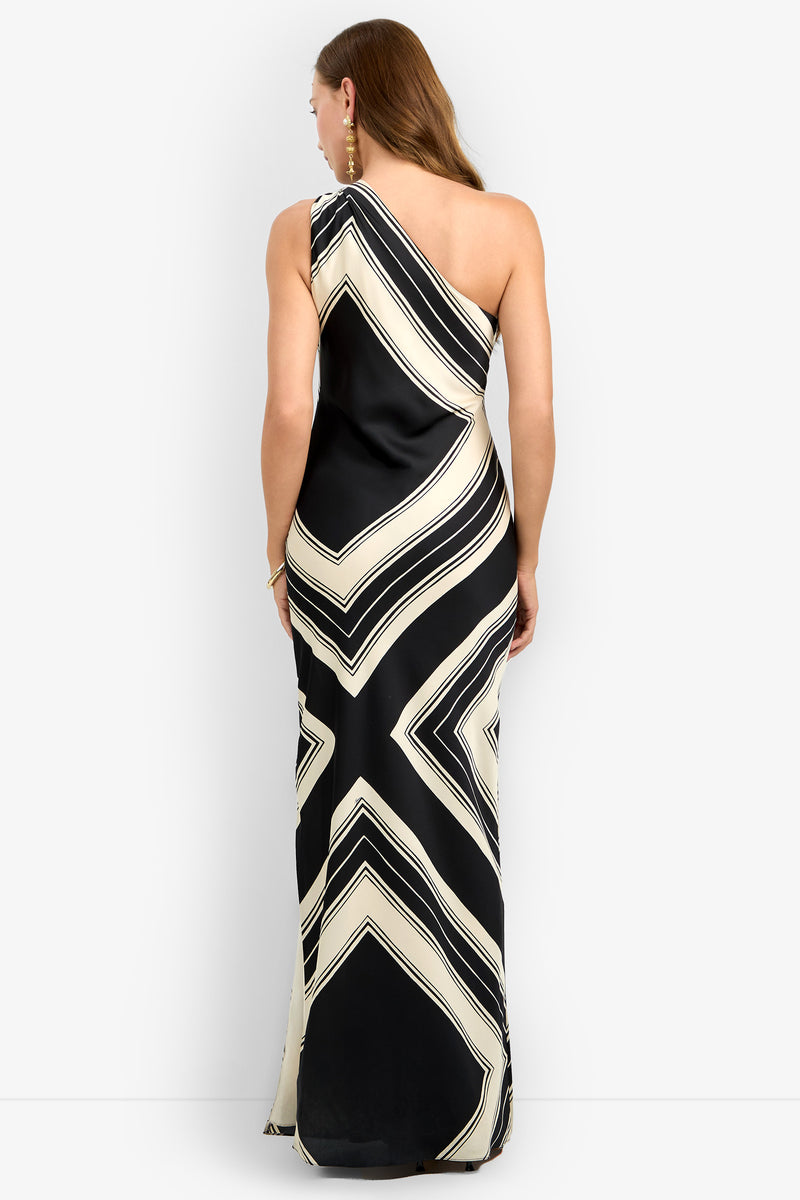 A woman models the CHIANTI DRESS, a one-shoulder, floor-length black gown with striking cream and white geometric patterns, posing to showcase the back against a plain backdrop.