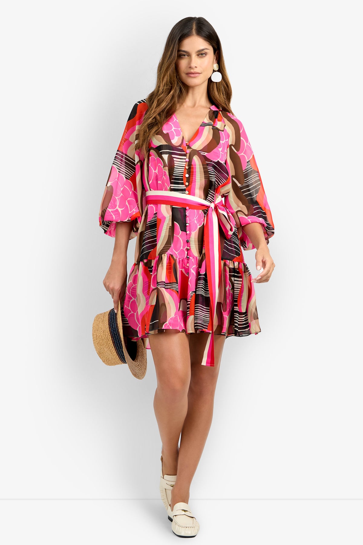 Woman wearing the AMATAS DRESS with pink, red, and brown patterns and a tied waist belt, paired with white loafers and holding a straw hat. She stands against a white background.