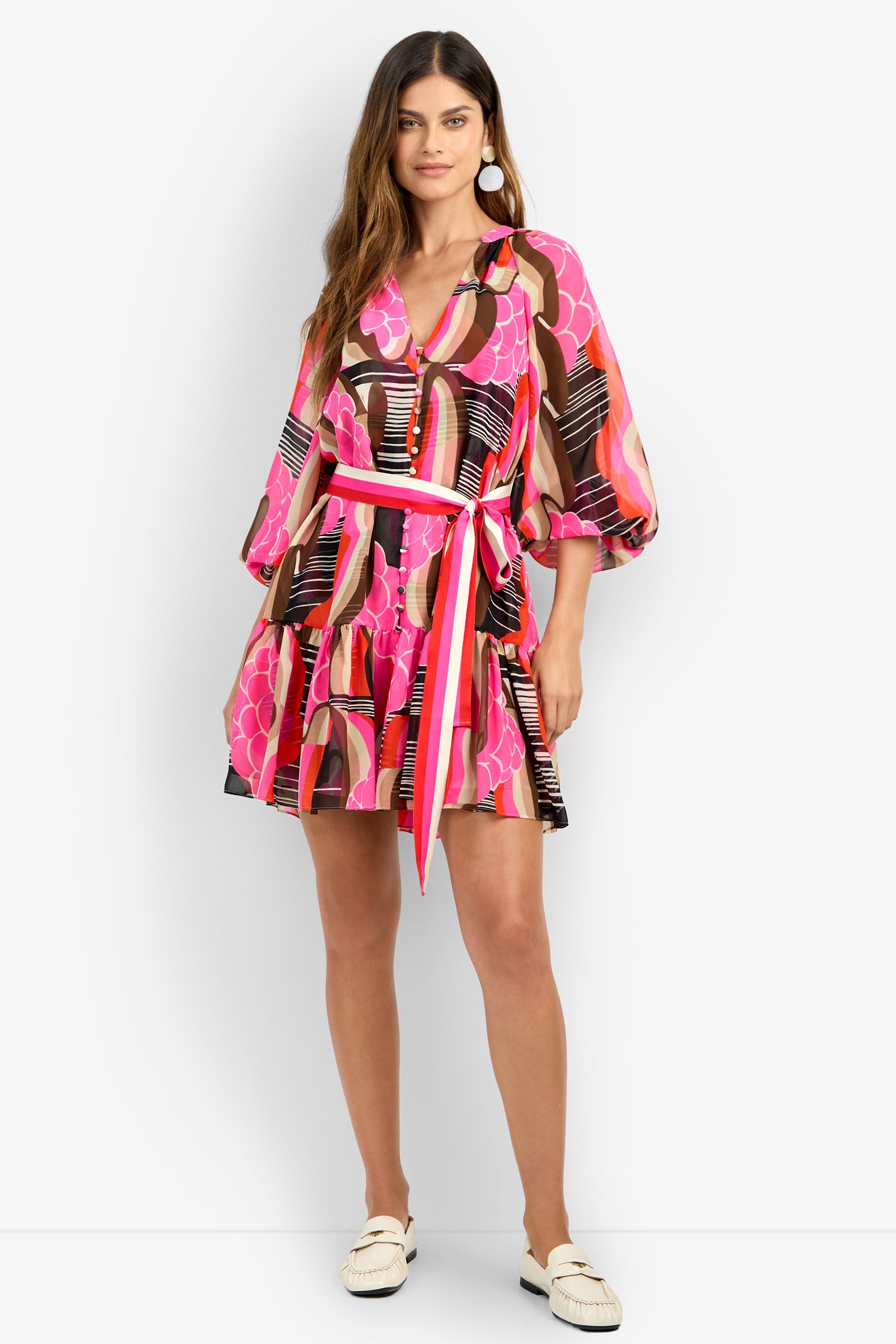 A woman stands and smiles in the AMATAS DRESS, a colorful V-neck dress with pink, orange, and black patterns, puffy sleeves, and a tied waist belt. She pairs it with white shoes and large round earrings.