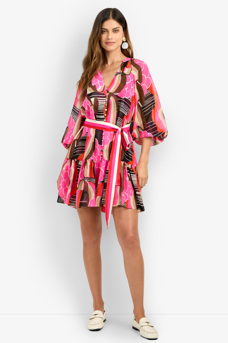 A woman stands and smiles in the AMATAS DRESS, a colorful V-neck dress with pink, orange, and black patterns, puffy sleeves, and a tied waist belt. She pairs it with white shoes and large round earrings.