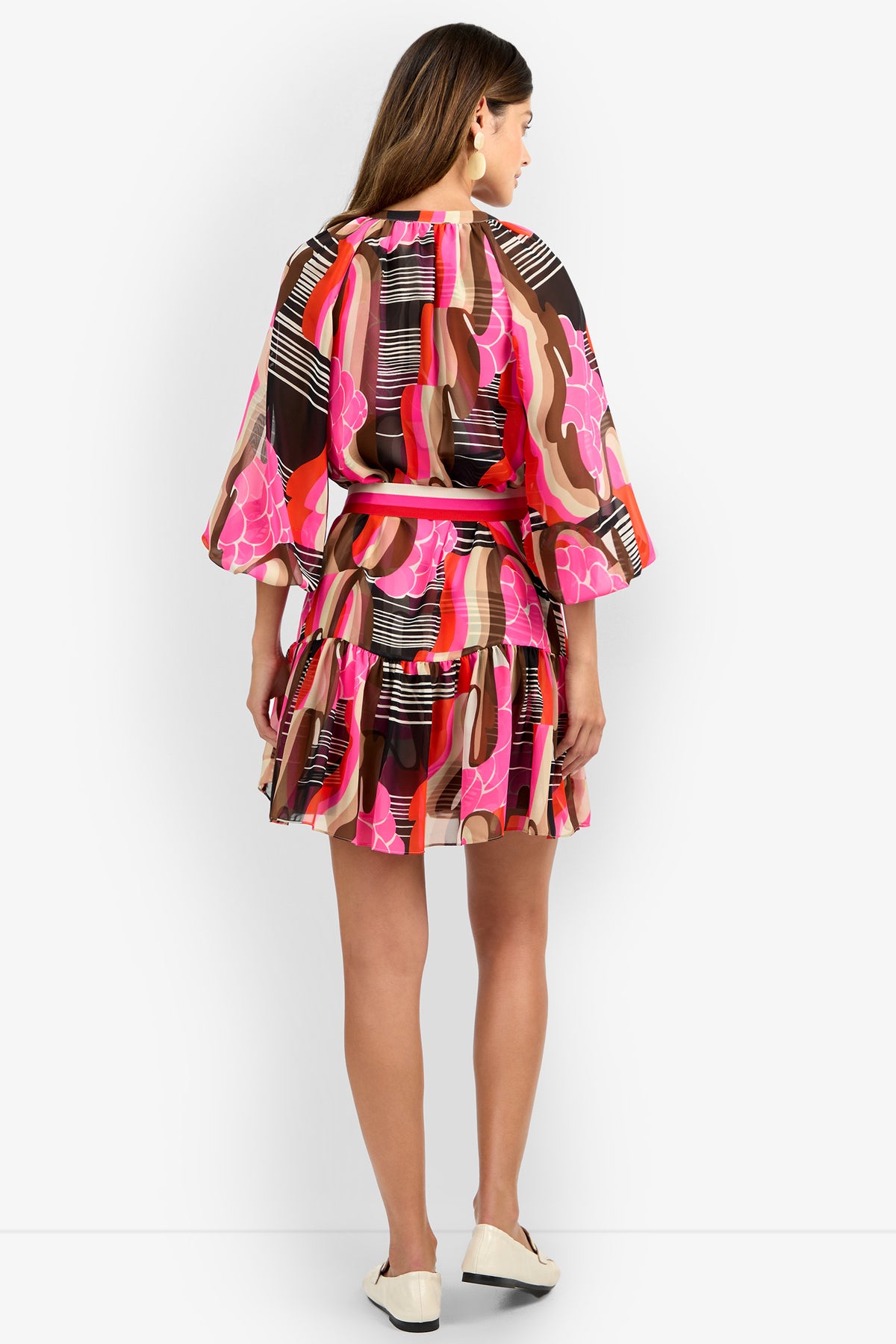A woman with light brown hair faces away, wearing the AMATAS DRESS featuring colorful pink, red, black, and brown abstract patterns, loose sleeves, a ruffled skirt, and white shoes.