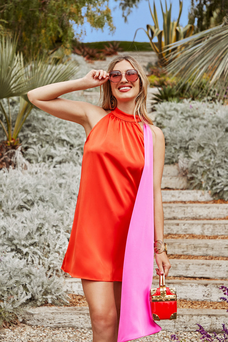 Wearing the VANNI DRESS, a woman stands outdoors, smiling and holding pink sunglasses. The sleeveless orange dress features a long pink sash, and she carries a small red handbag amidst lush green foliage and stone steps.