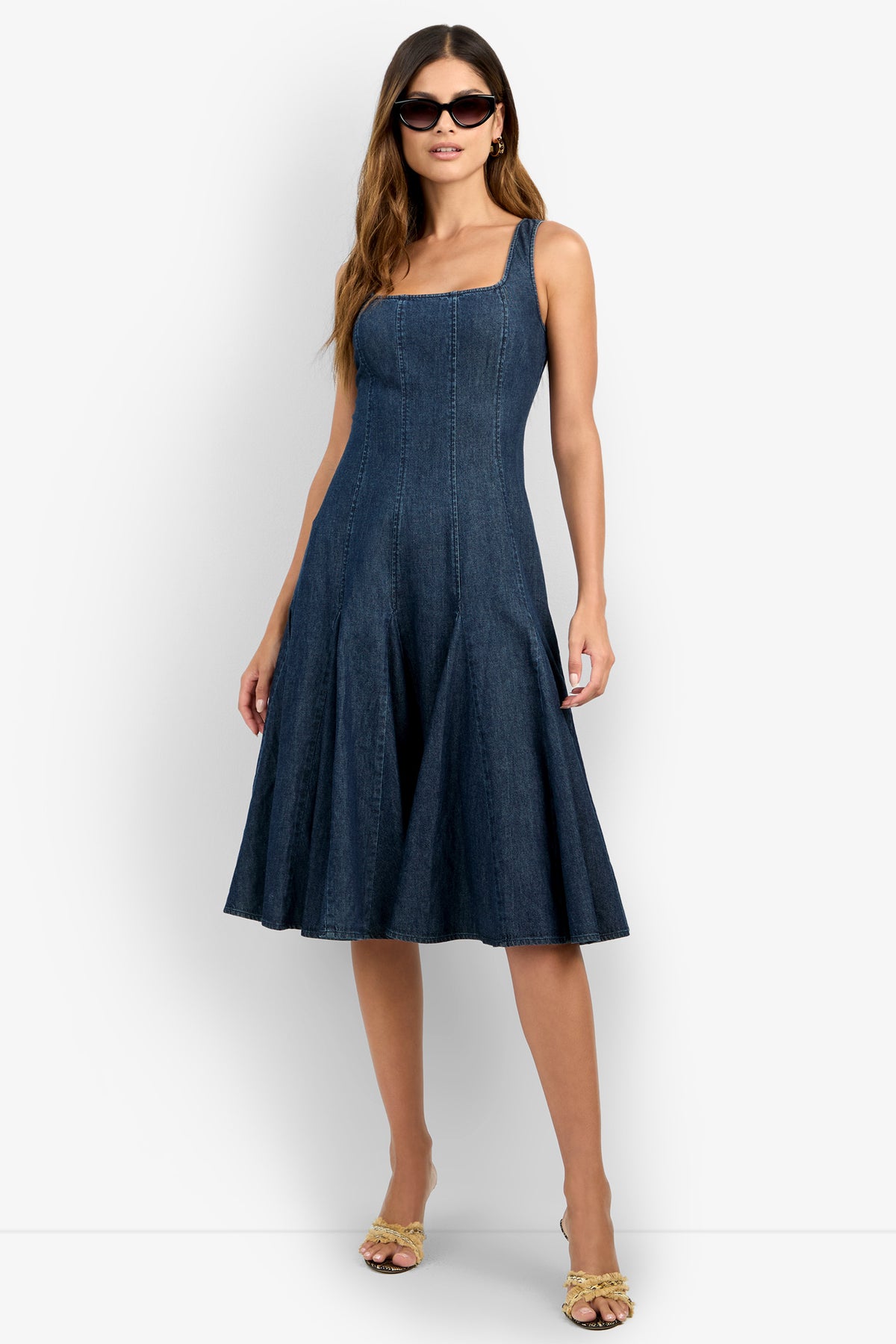 A woman poses against a white background wearing the CASIMA DRESS—a sleeveless, dark blue denim fit-and-flare dress with a square neckline and knee-length hem—paired with sunglasses and open-toe heels, one hand on her hip.