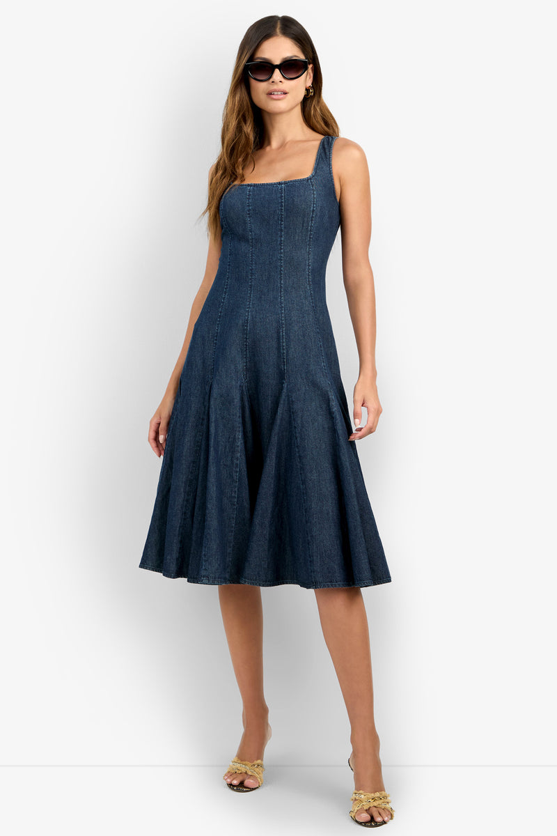 A woman poses against a white background wearing the CASIMA DRESS—a sleeveless, dark blue denim fit-and-flare dress with a square neckline and knee-length hem—paired with sunglasses and open-toe heels, one hand on her hip.