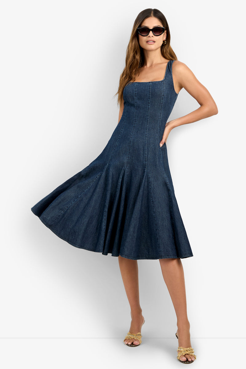 Woman models the CASIMA DRESS, a sleeveless dark blue denim piece with a flared skirt. She accessorizes with sunglasses and strappy sandals, her long wavy hair flowing against a plain white background.