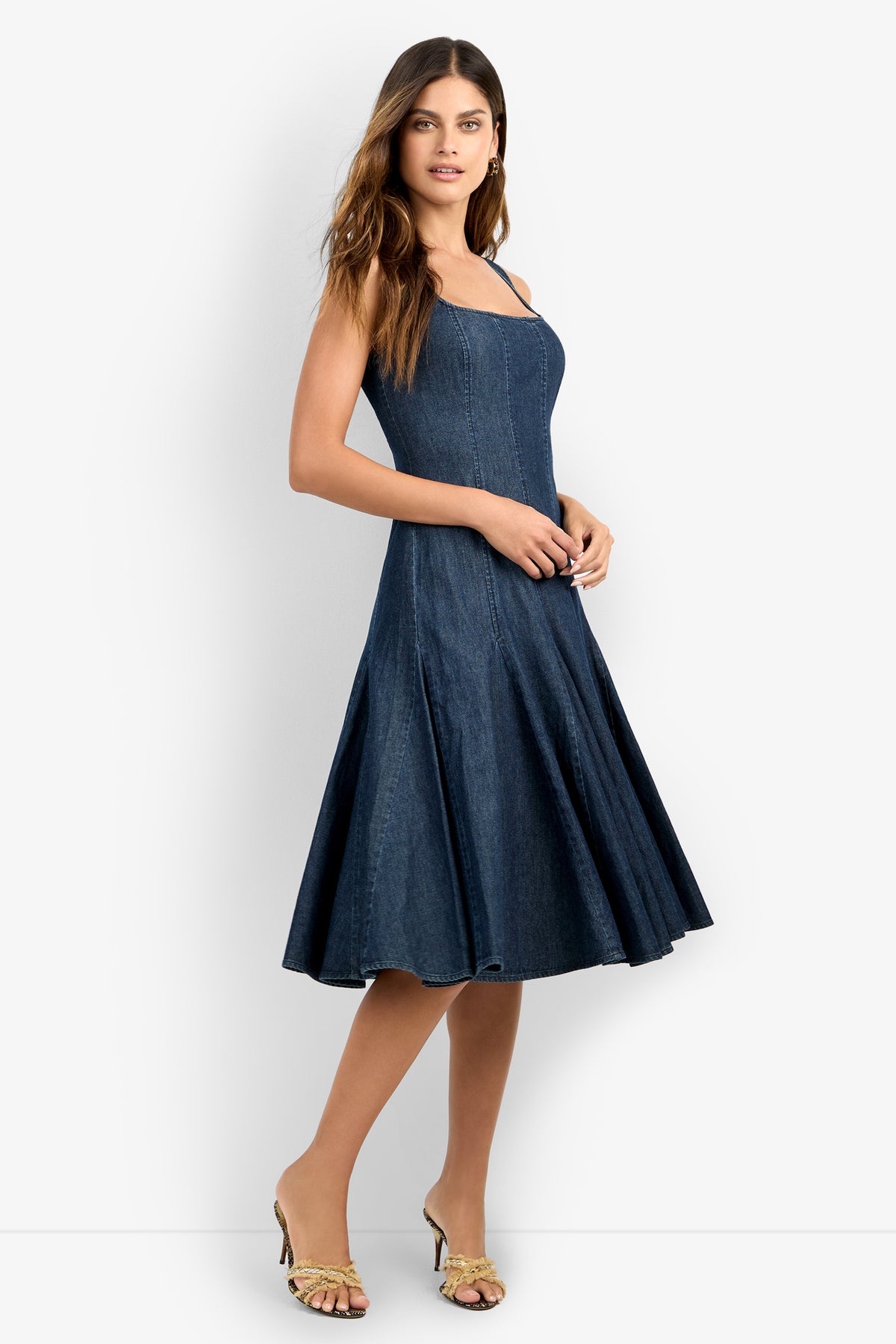 A woman with long brown hair stands sideways in heeled sandals against a white background, wearing the CASIMA DRESS—a sleeveless dark blue denim midi dress featuring a fitted bodice and flared skirt.